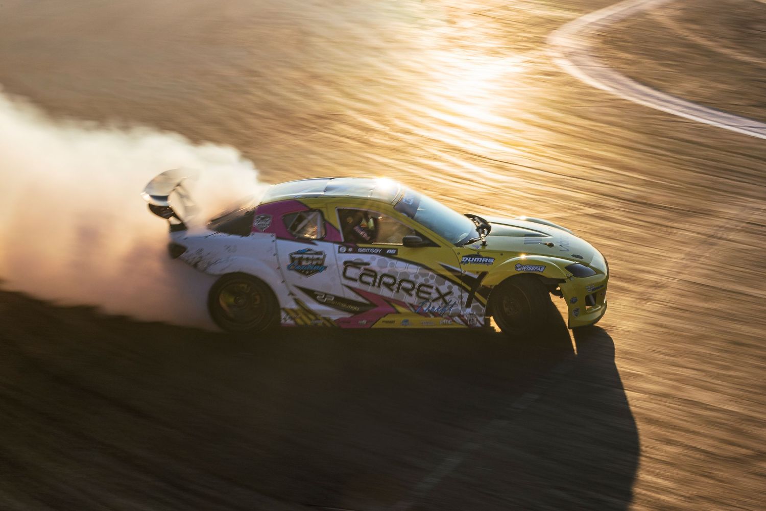 Aerial view of a yellow and pink drift car sliding on a dusty track at sunset, casting a long shadow and kicking up smoke.