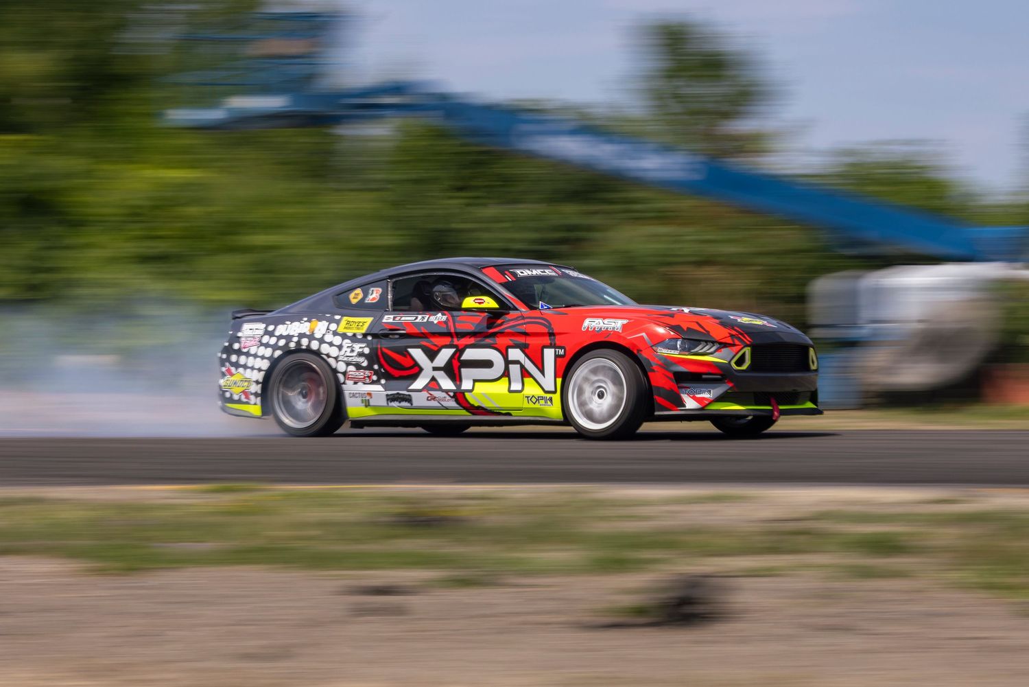 Red, yellow and black XPN drift car at speed on an asphalt track with motion blur and light smoke trailing behind.
