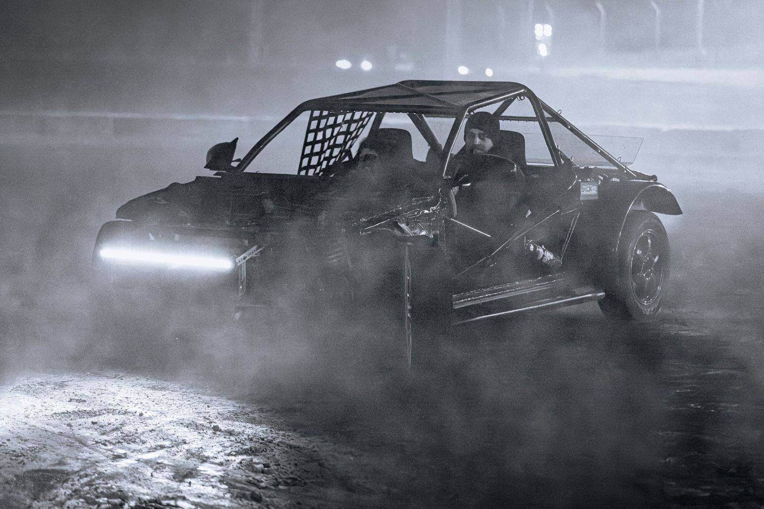 Black and white image of an open-frame off-road drift buggy with a roll cage and light bar moving through mist.