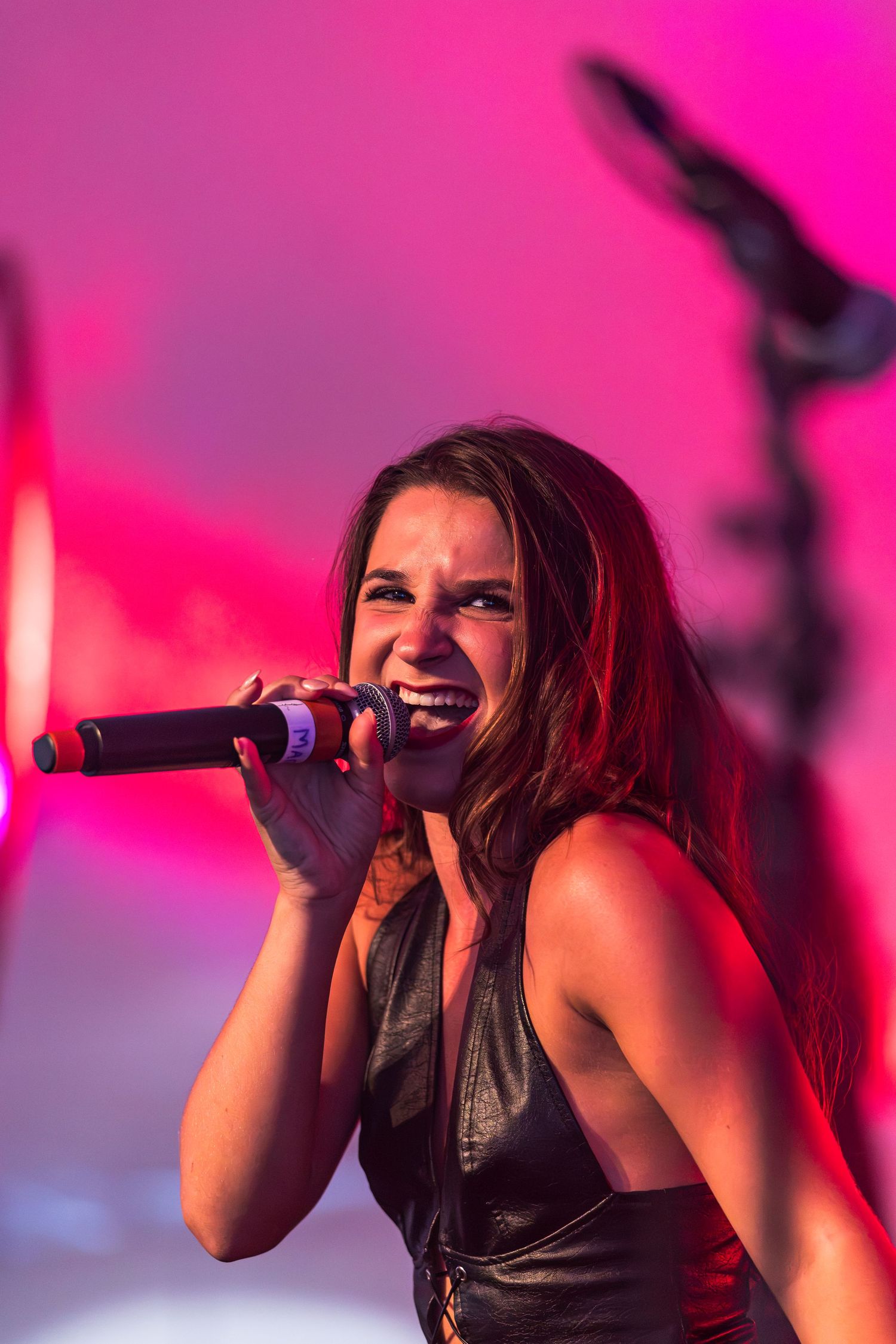 Female singer in a black leather top sings passionately into a handheld microphone on stage against a vivid pink and purple backdrop.