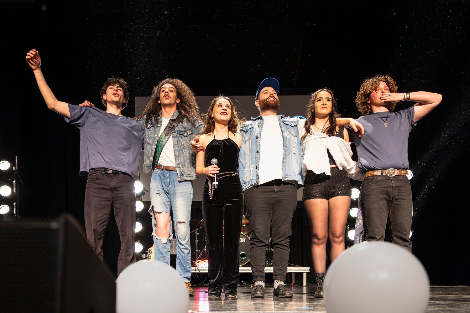 Six performers stand arm in arm taking a bow on stage, with balloons in the foreground and stage lights behind.