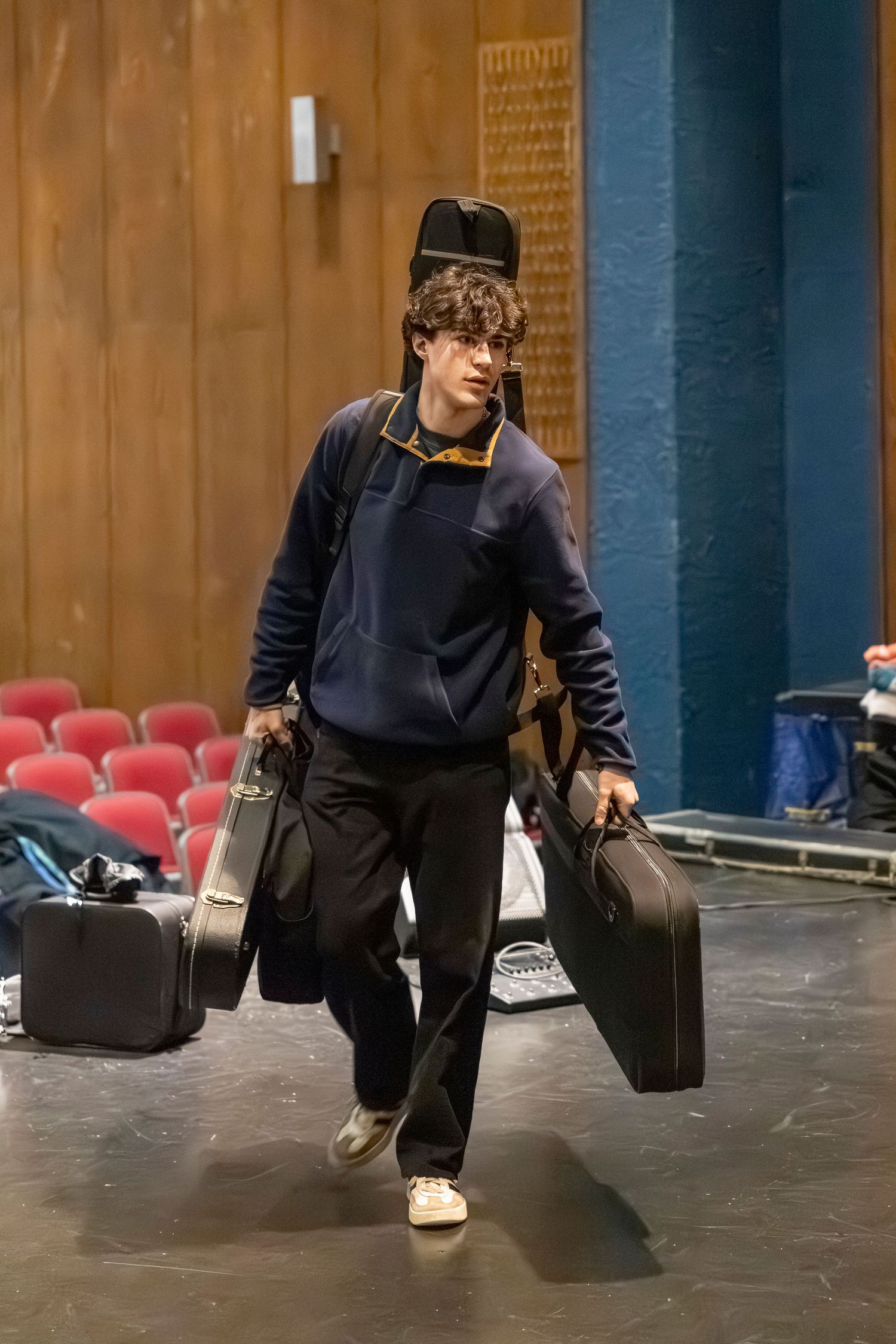 A young man in a navy sweater carries several instrument cases across a stage with red theater seats behind him.