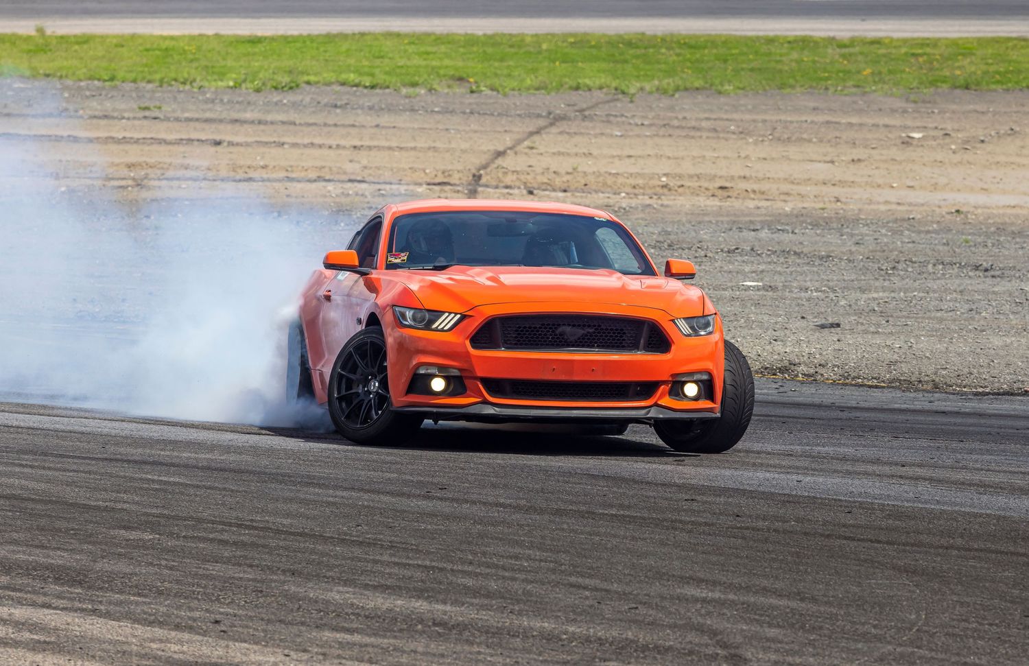 Orange Ford Mustang drifting on an asphalt track with tire smoke trailing from its rear wheels.