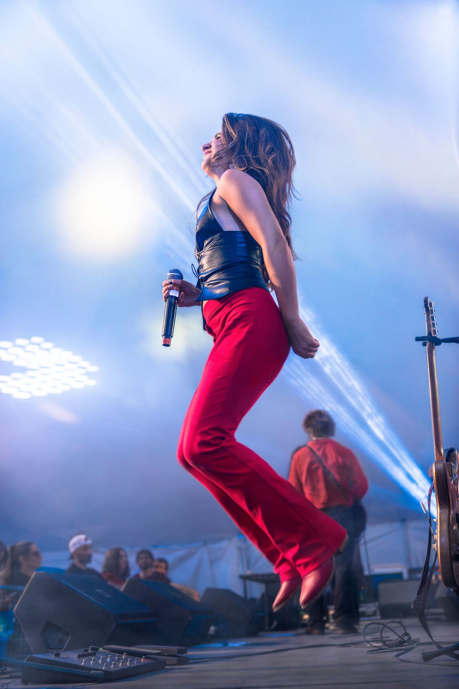Female singer in red pants and black leather top leaps mid-air holding a microphone on a stage lit by blue lights while the crowd watches.