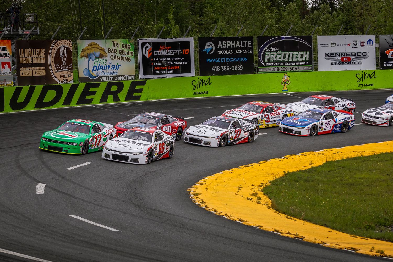 Pack of stock cars racing closely through a curve on a paved circuit lined with sponsor banners.