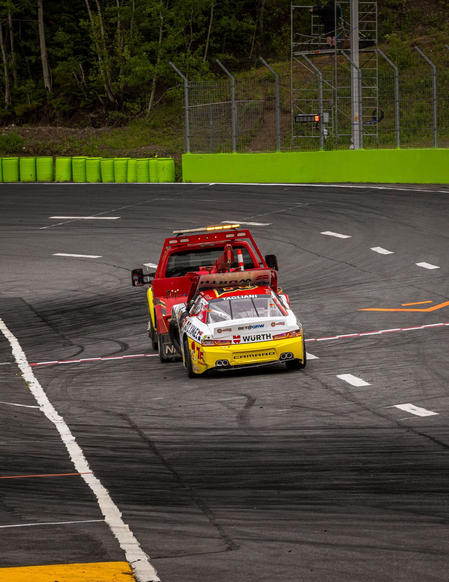 A red tow truck pulls a damaged yellow and white stock car along an asphalt race track beside green barriers.