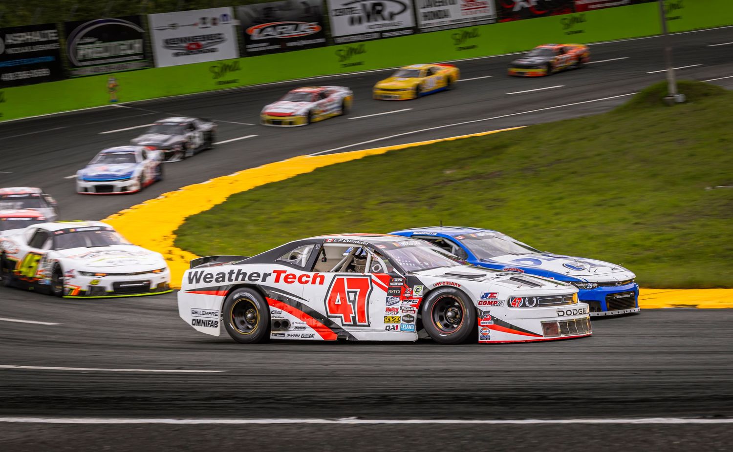 A WeatherTech-sponsored number 47 stock car races on an asphalt track, chasing another car with a pack of racers ahead.