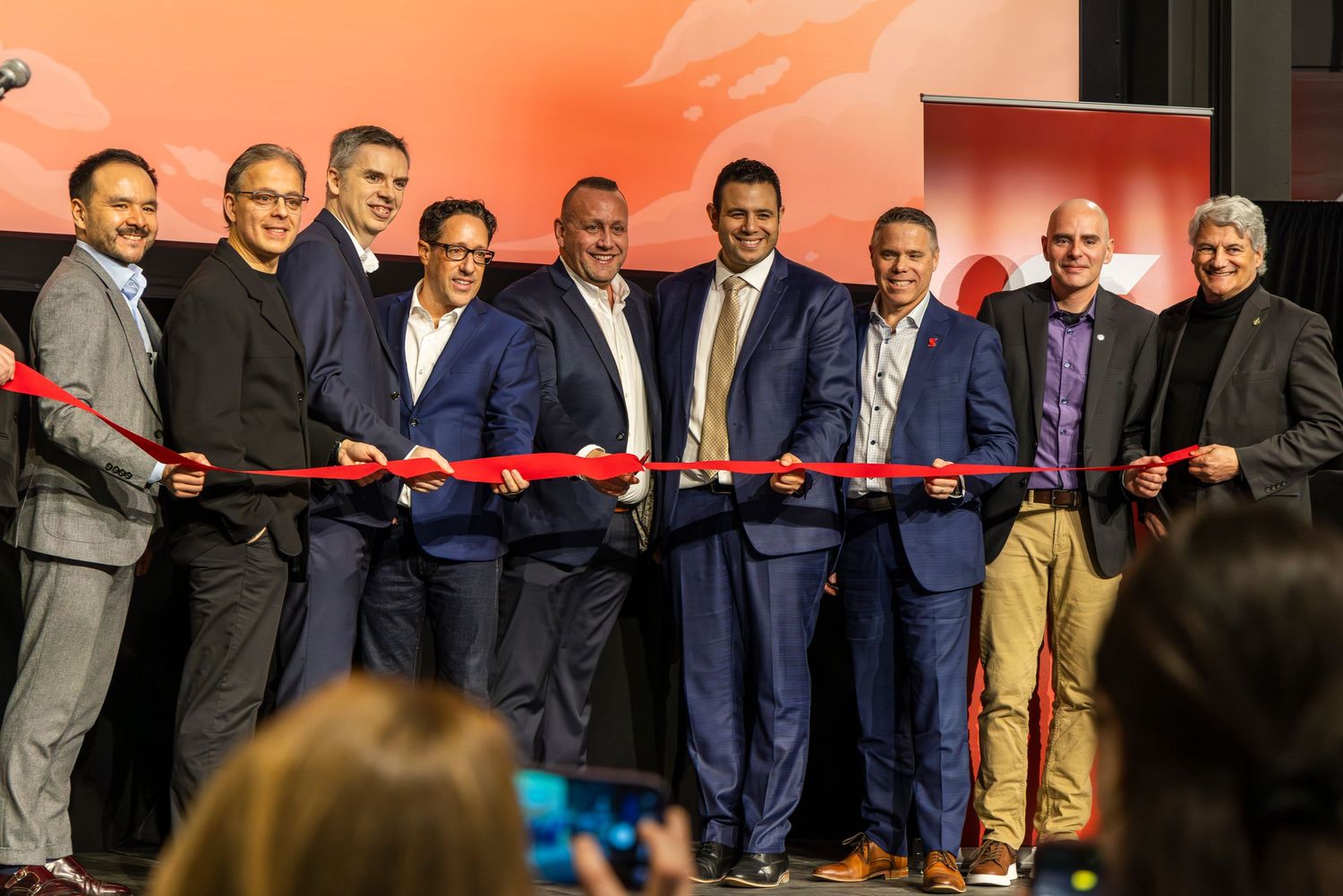 Seven men in business attire stand in a row holding a red ribbon during a ribbon-cutting ceremony at a corporate event.