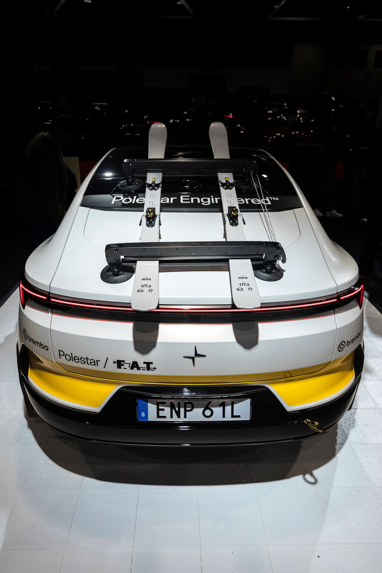 Rear view of a white Polestar electric car with yellow accents and skis mounted on the roof, displayed at an indoor auto show.