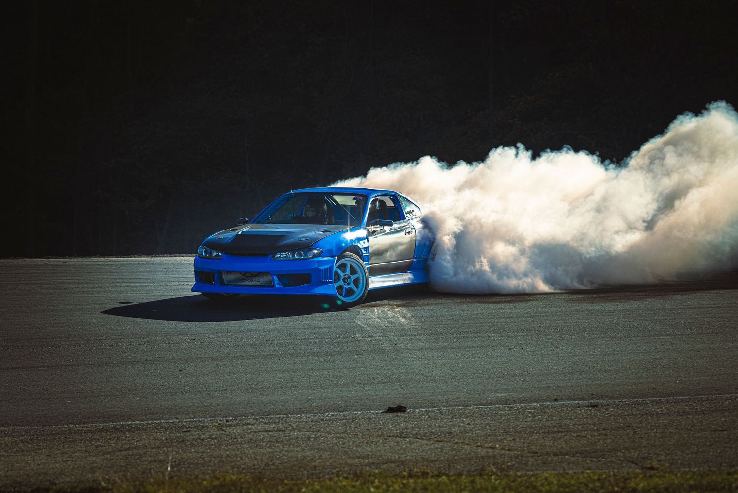 Blue drift car carving a sharp slide on a track while a huge cloud of tire smoke rises behind it.