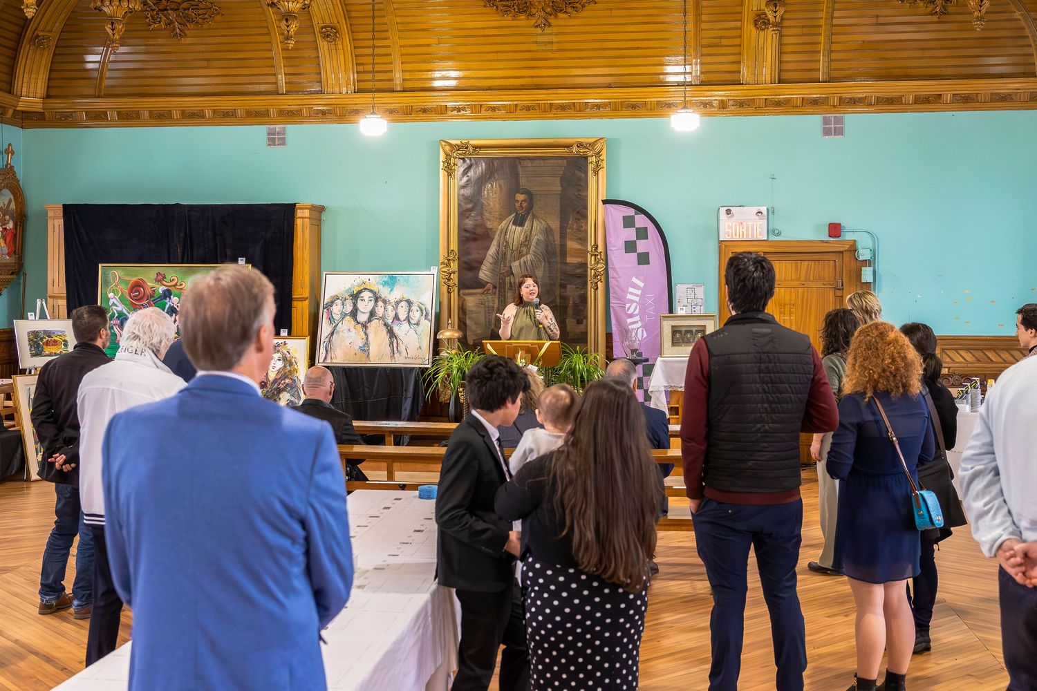 Visitors mingle in a hall with teal walls and wood trim, viewing framed artwork and displays at an exhibition.