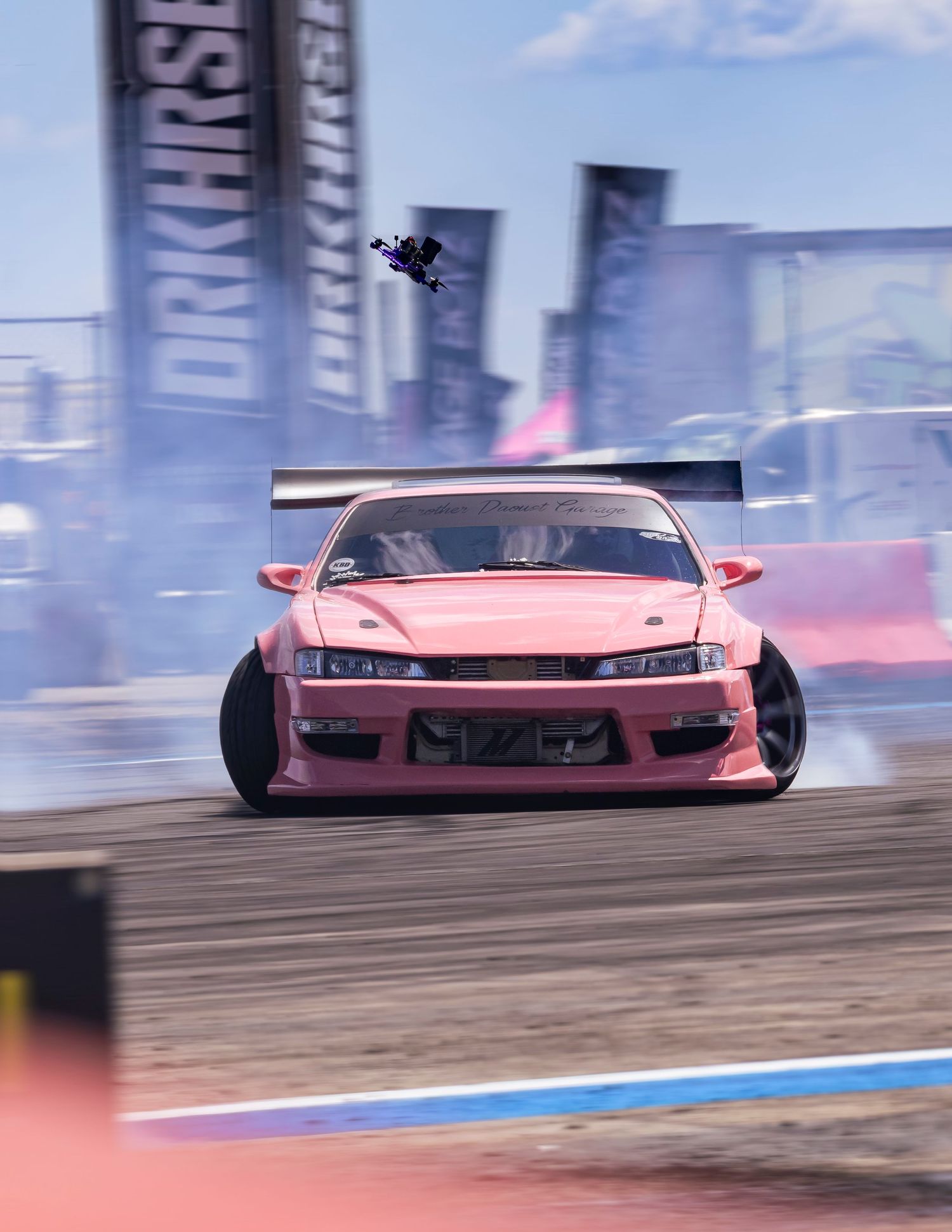 Pink drift car sliding sideways through smoke at a drift event with sponsor banners and a drone flying overhead.