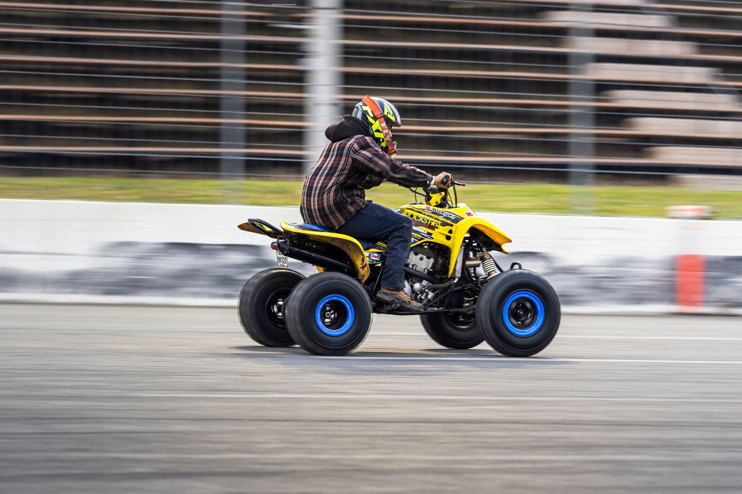 Rider in a colorful helmet speeds past on a yellow quad bike with blue wheels along a race track.