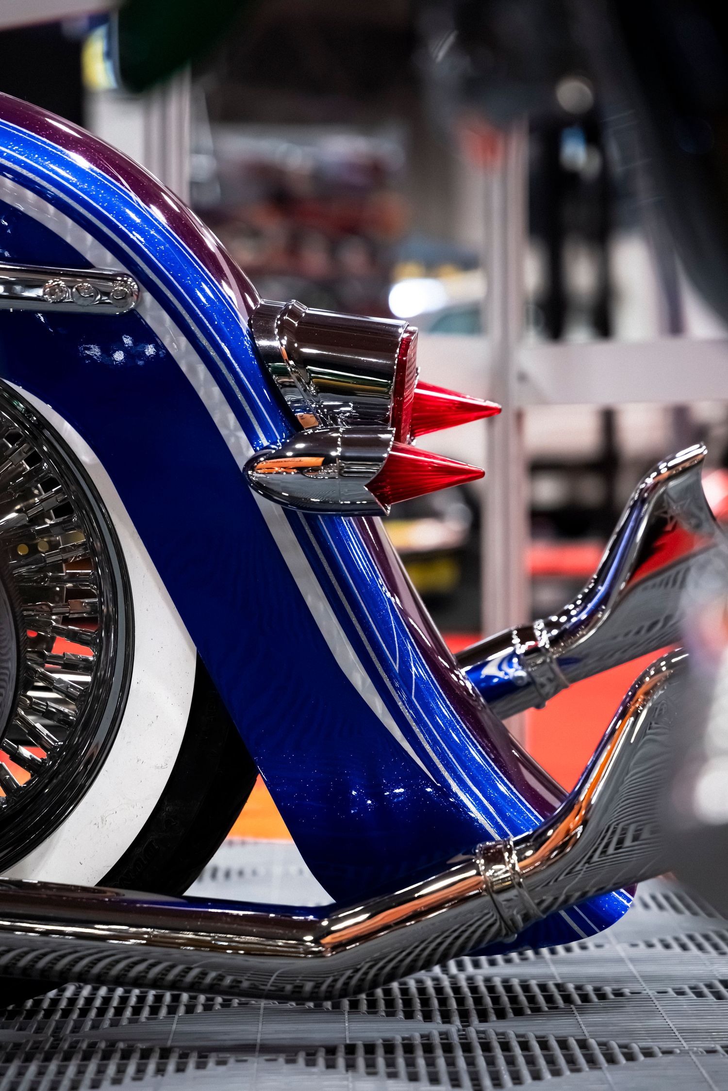 Close-up of a custom motorcycle with a glossy blue painted fender, chrome exhaust pipes, red accents and a whitewall wire wheel.