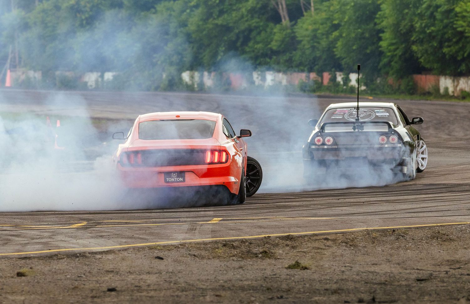 Two drift cars, an orange Ford Mustang and a dark sports car, sliding side by side on a racetrack in clouds of tire smoke.