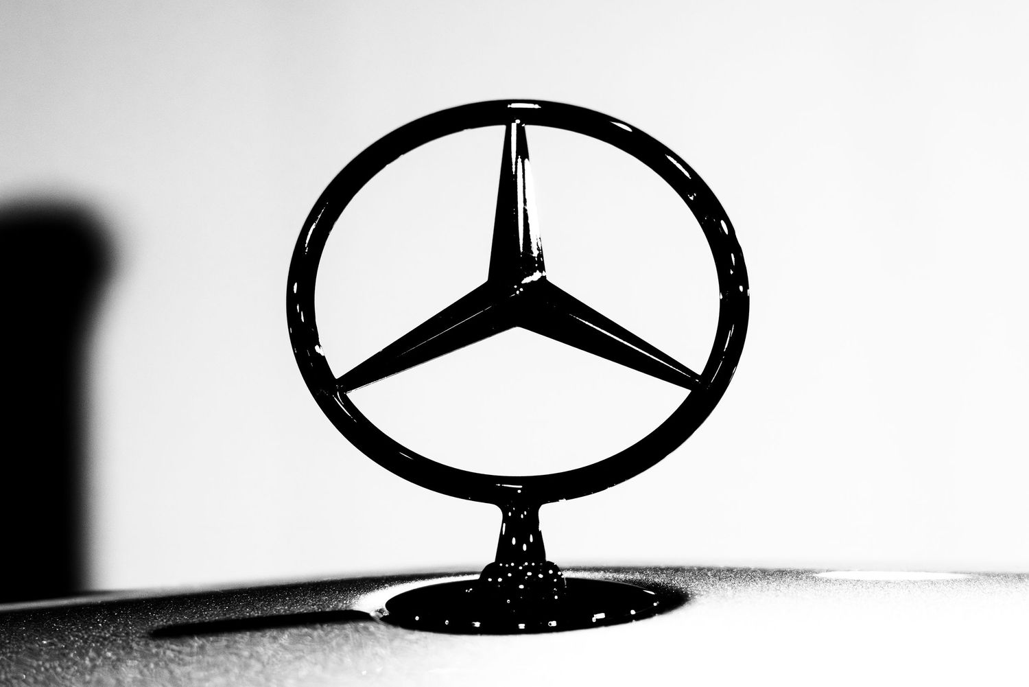 Close-up black and white photo of a Mercedes-Benz three-pointed star hood ornament with water droplets on the hood.
