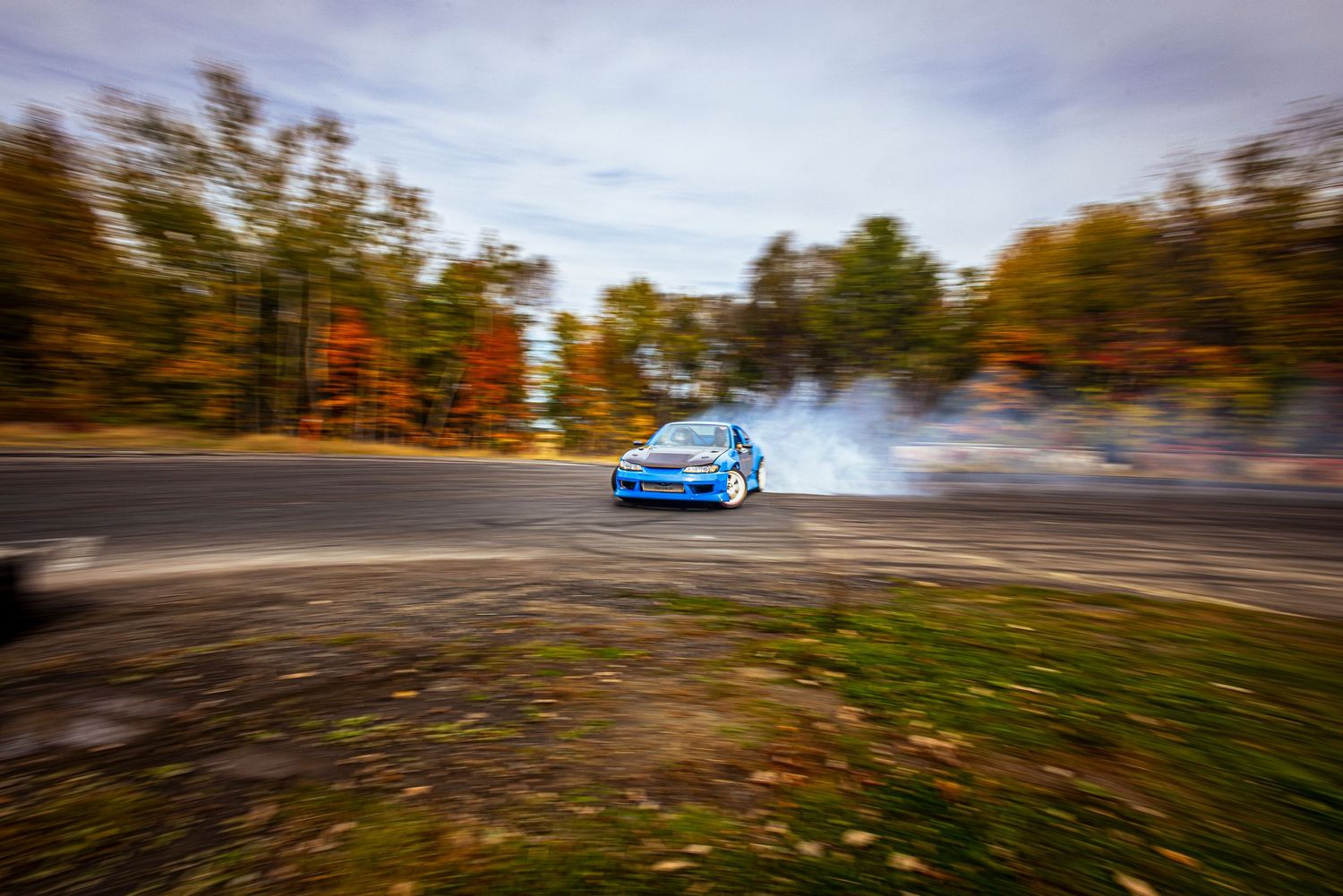 A blue car drifts around a curve on an asphalt track, trailing tire smoke past colorful autumn trees.