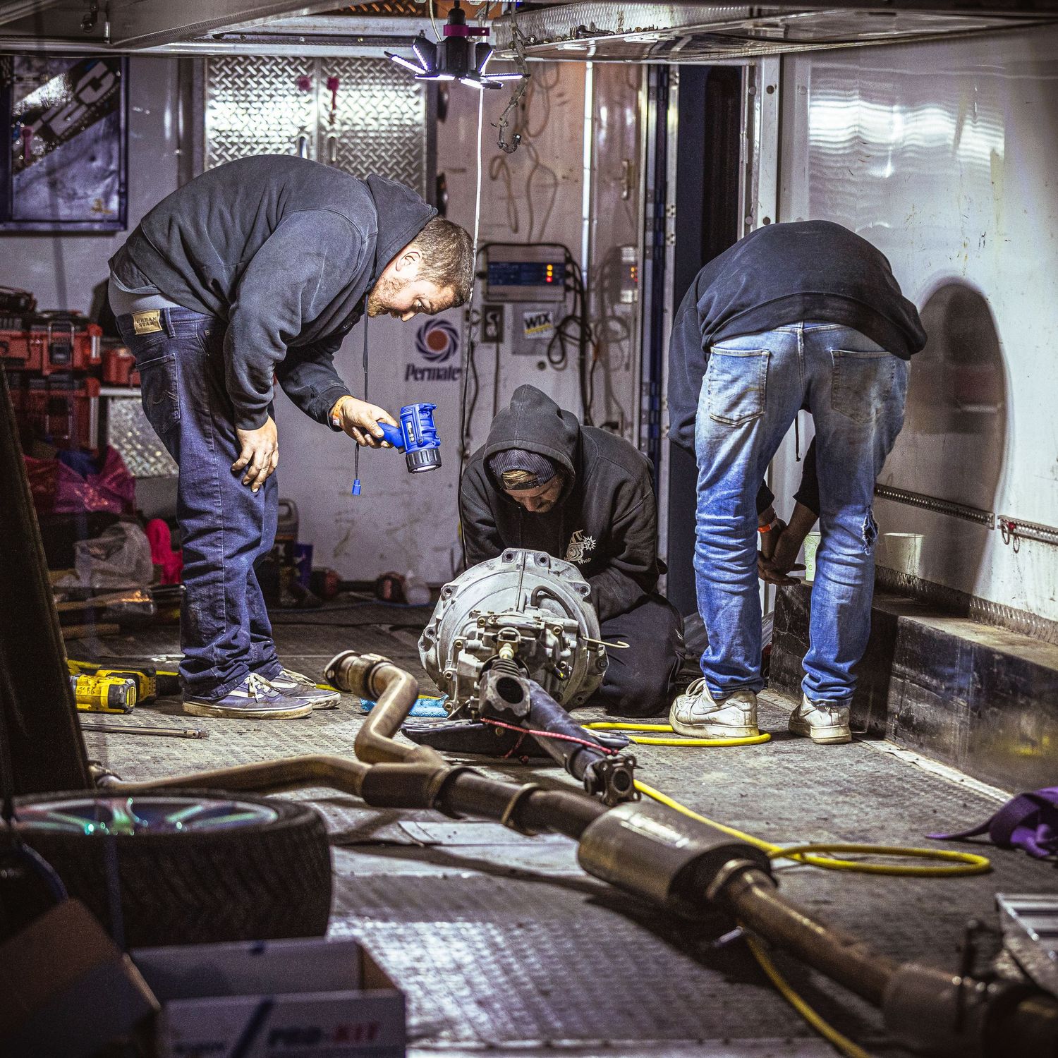 Mechanics work on an exposed drivetrain and exhaust parts inside a race trailer garage with tools and equipment around.