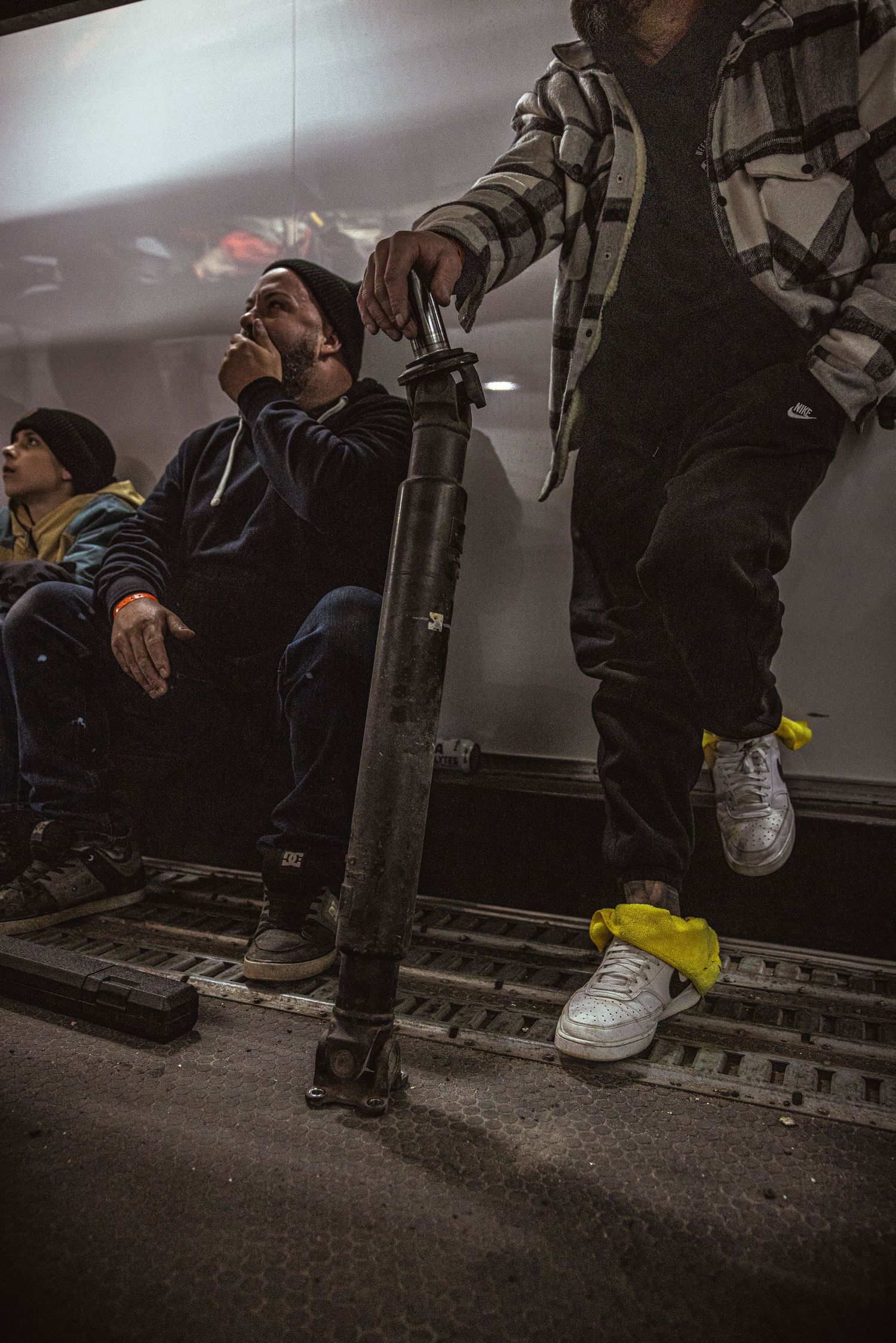 Two men sit and another stands with a foot on a metal rail, holding a long black shock absorber inside a trailer.