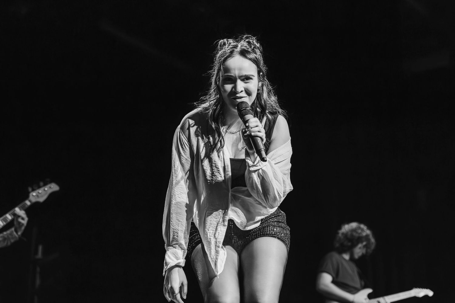 Black and white photo of a singer in an open shirt and shorts leaning forward with a microphone as band members play behind.