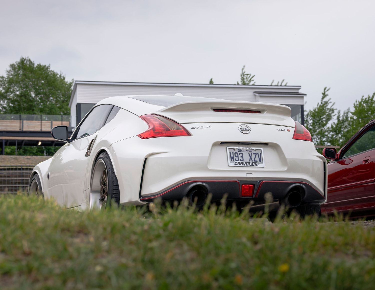 Rear view of a white Nissan 370Z Nismo sports car with a spoiler and dual exhaust parked on grass beside a red car.