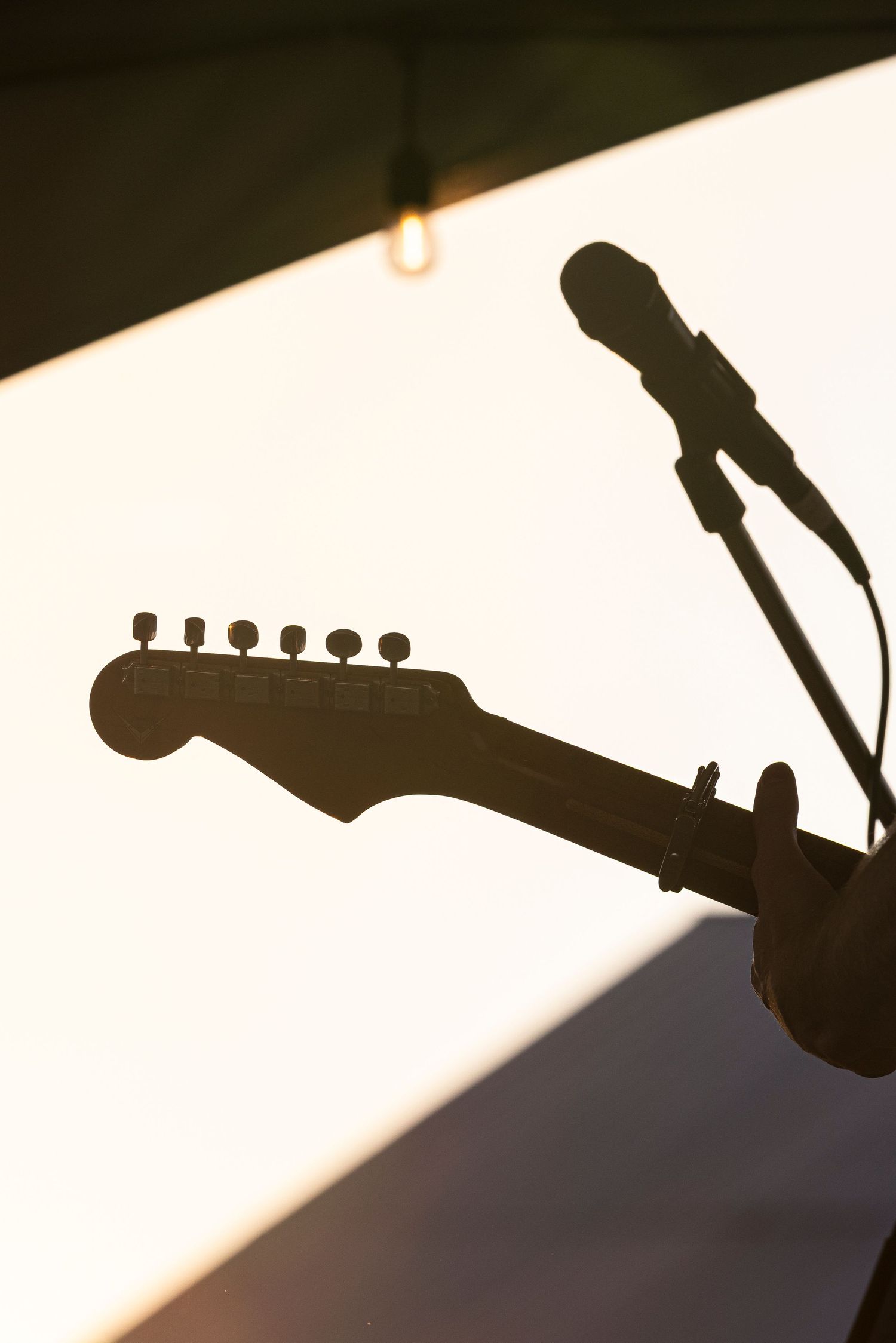 Silhouette of an electric guitar headstock and microphone stand against a bright sunset sky with a hanging string light overhead.