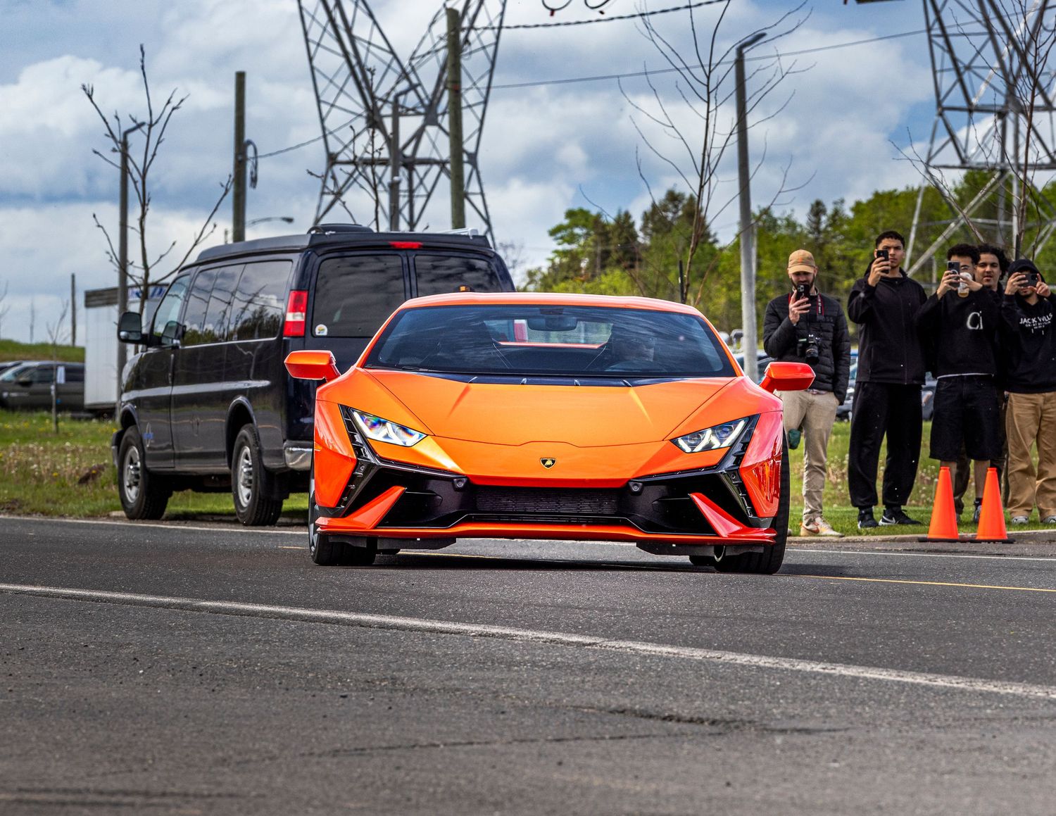 Orange Lamborghini Huracán Tecnica supercar driving on a track with spectators and orange traffic cones near a black SUV in the background.