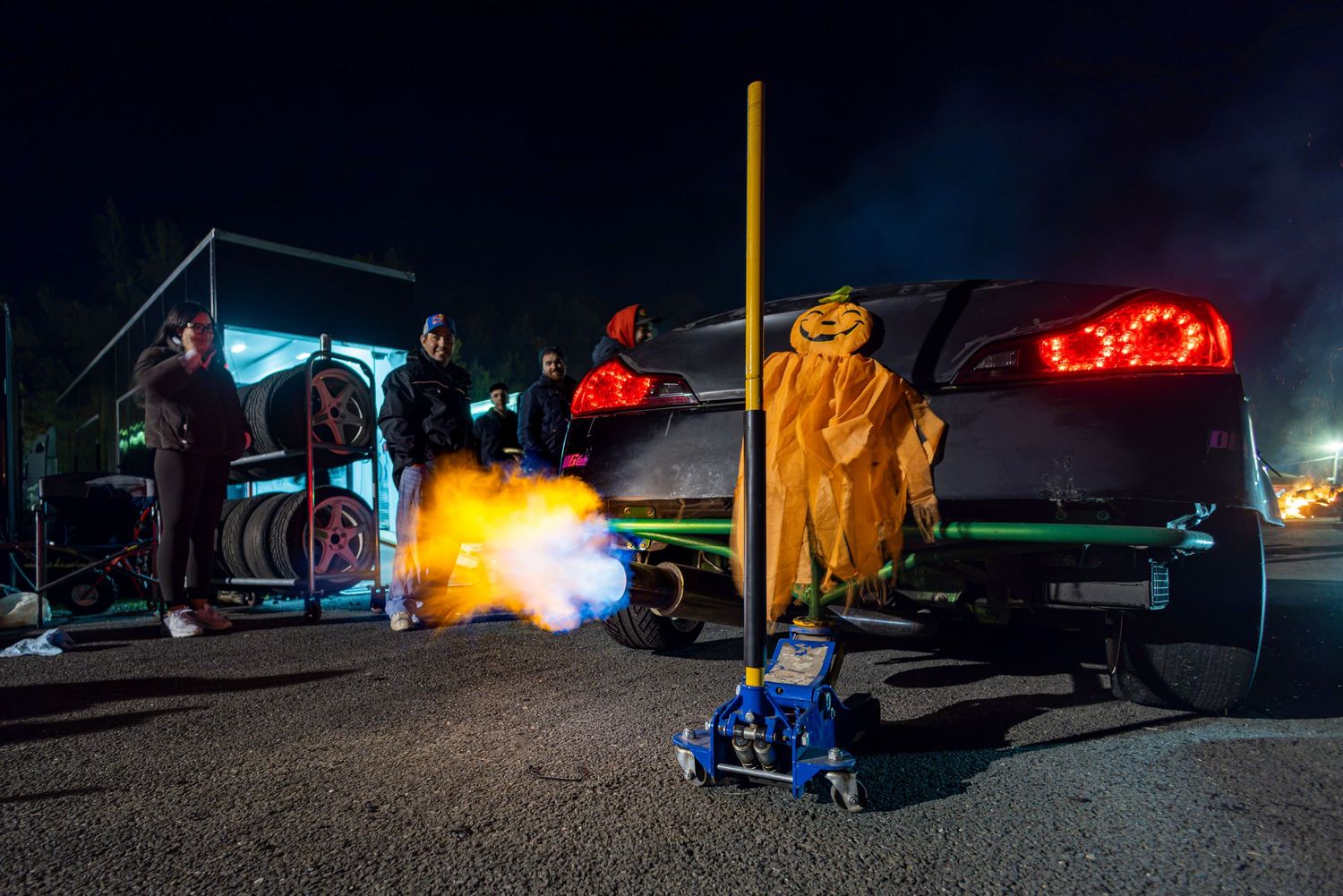 Spectators stand near a dark car at night as a flame bursts from its exhaust beside a jack and a costumed pumpkin figure.