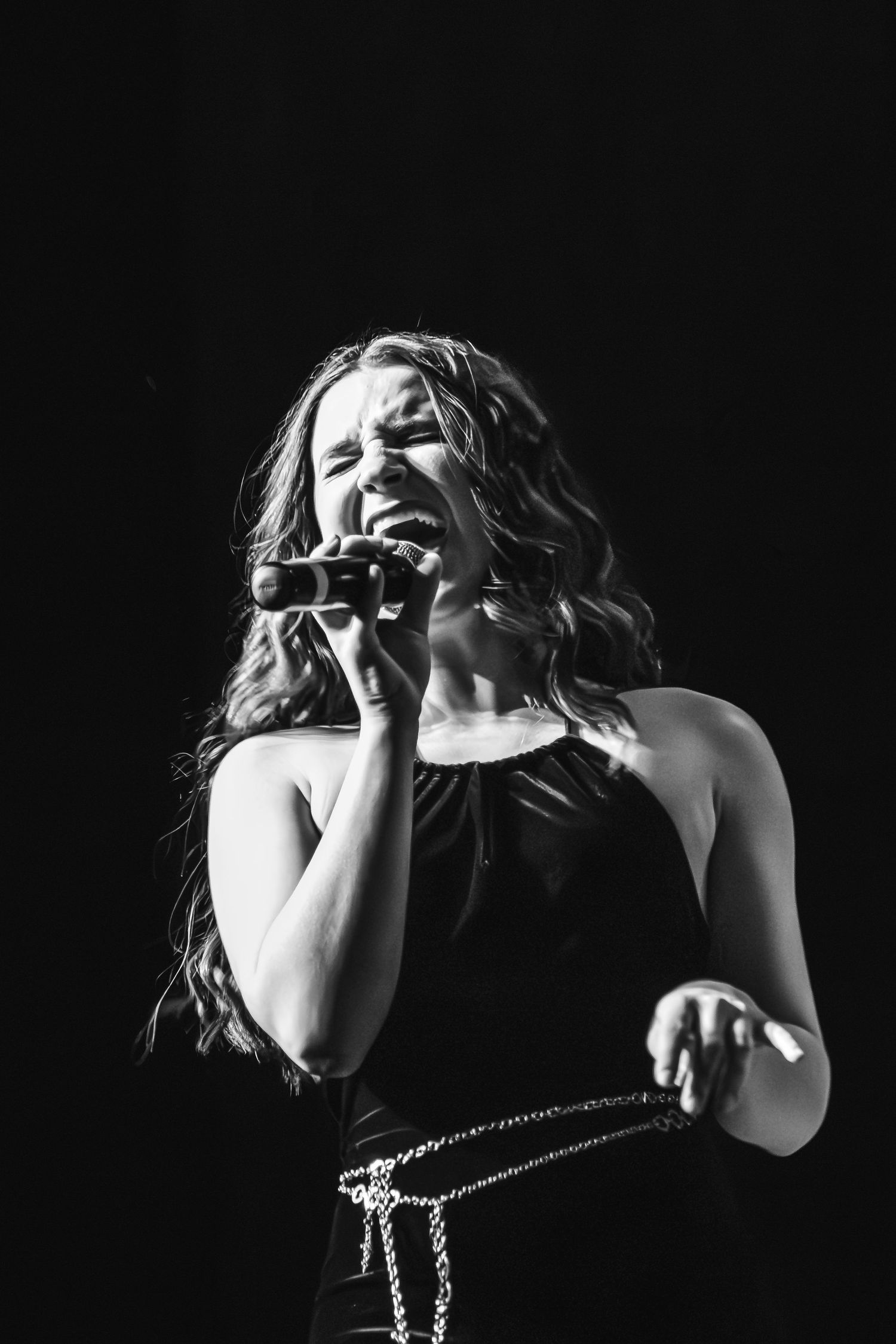 Black and white portrait of a female singer with long wavy hair belting into a microphone in a dark halter top.