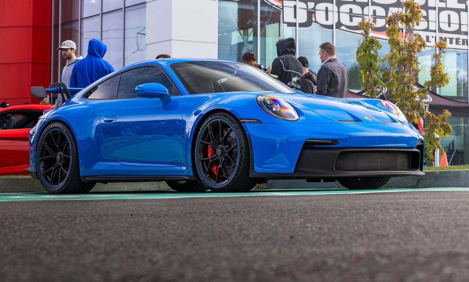 Blue Porsche 911 GT3 sports car with black wheels and red brake calipers parked at an outdoor car show near onlookers.