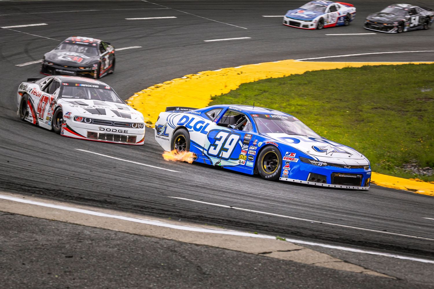 Stock cars including the blue number 99 racing closely on an asphalt oval track during a competitive race.