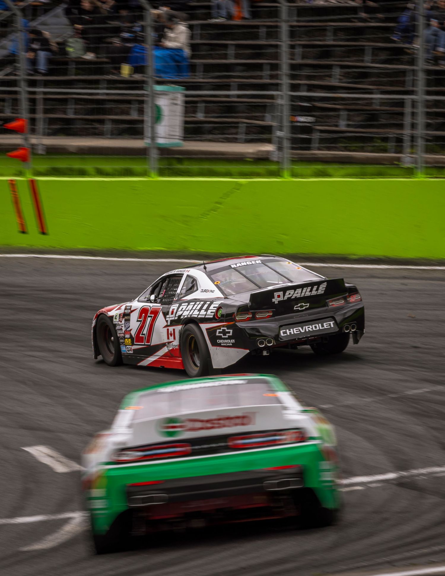 Two stock cars, a Chevrolet number 77 and a green car, racing on the track as spectators watch from the stands.