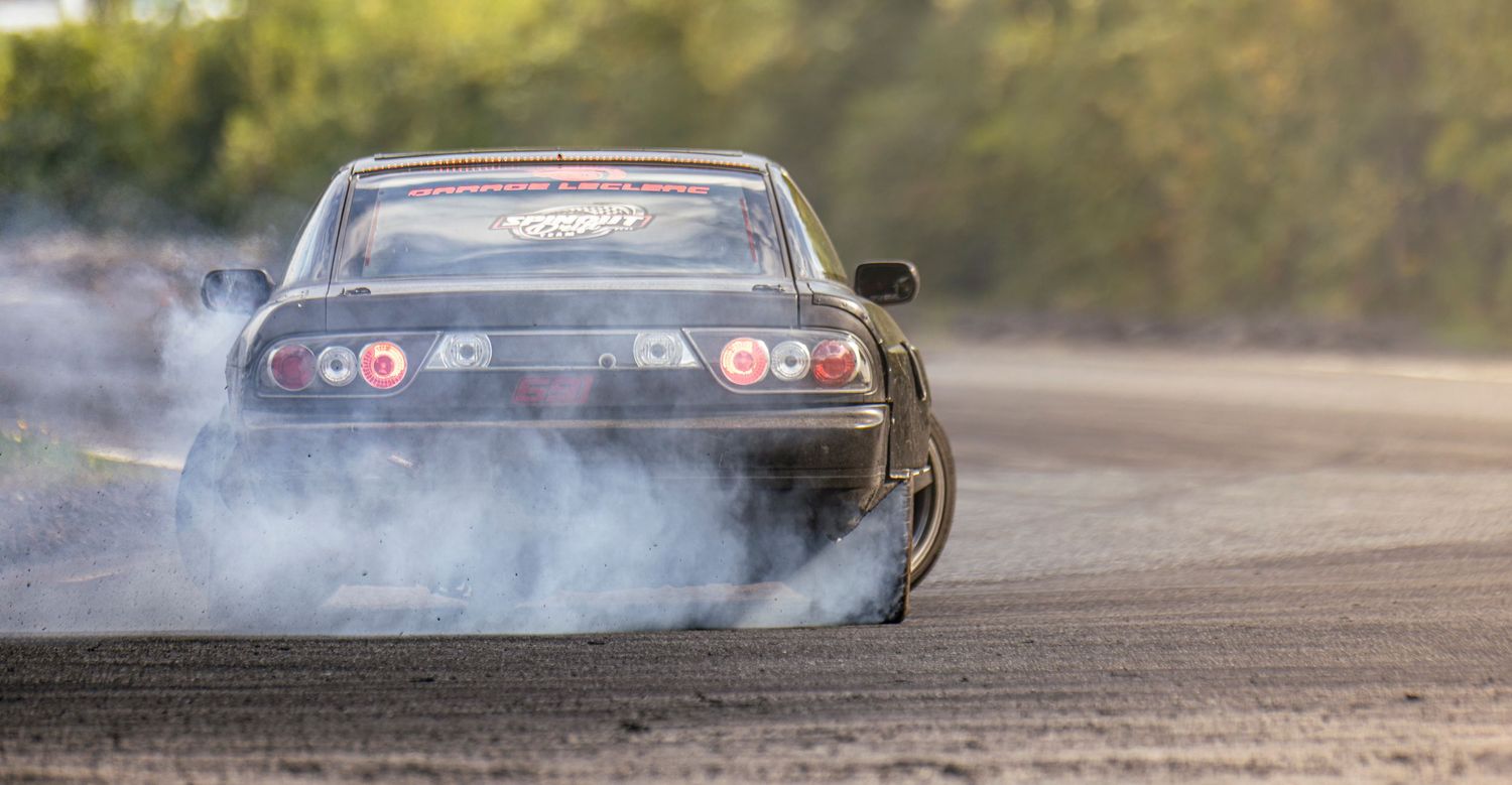Rear view of a drift car kicking up tire smoke while sliding around a curve on a track lined with trees.