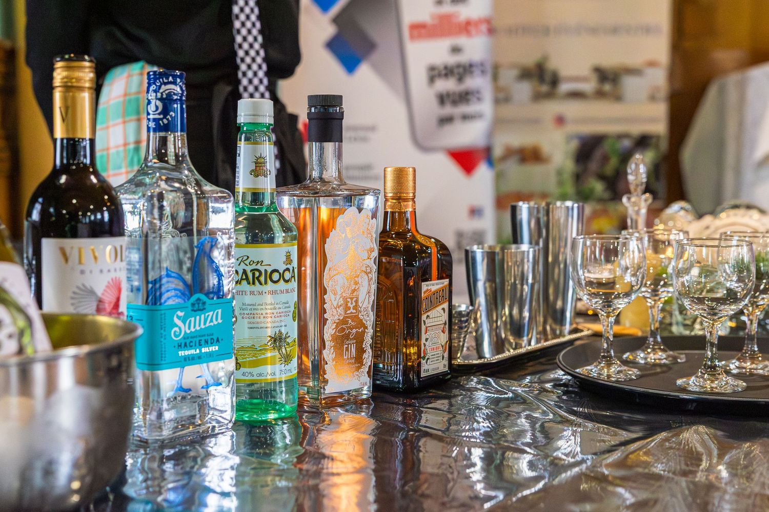 Bottles of wine, gin and liquor sit on a reflective bar counter beside metal shakers and empty cocktail glasses.