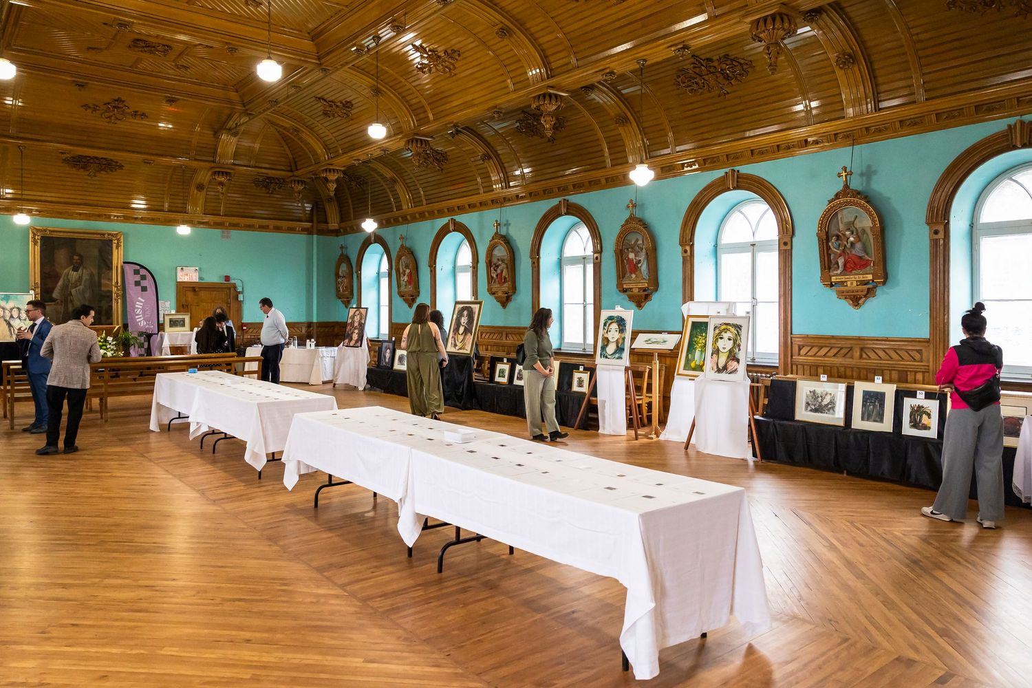 Visitors browse an exhibition in an ornate hall with teal walls, arched windows, coffered ceilings, white-draped tables and framed artwork.