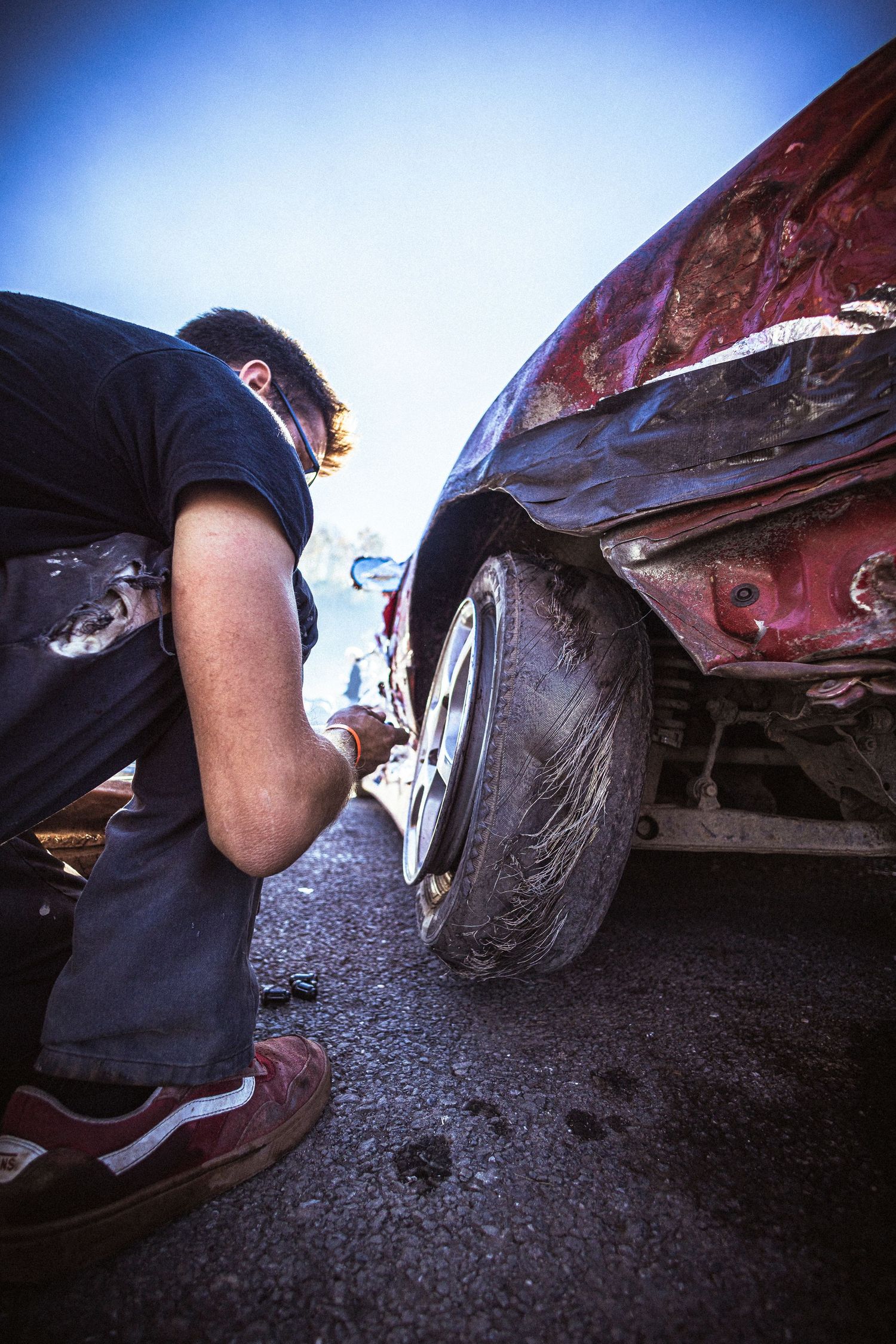 Mechanic crouches beside a damaged red car with a dented fender and worn tire, working on the wheel.