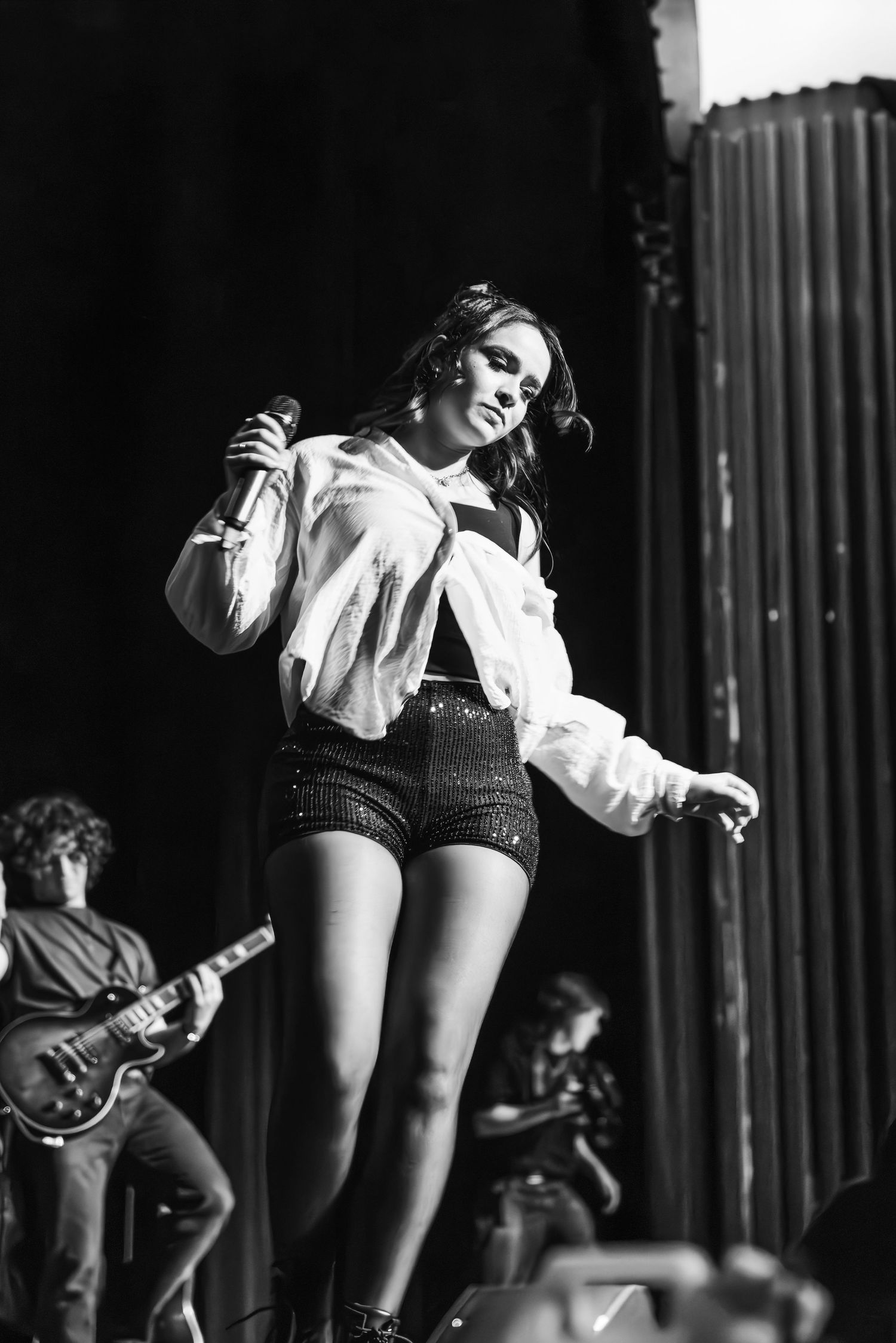 Black and white image of a singer in a white jacket and sparkly shorts holding a microphone on stage with a guitarist behind.