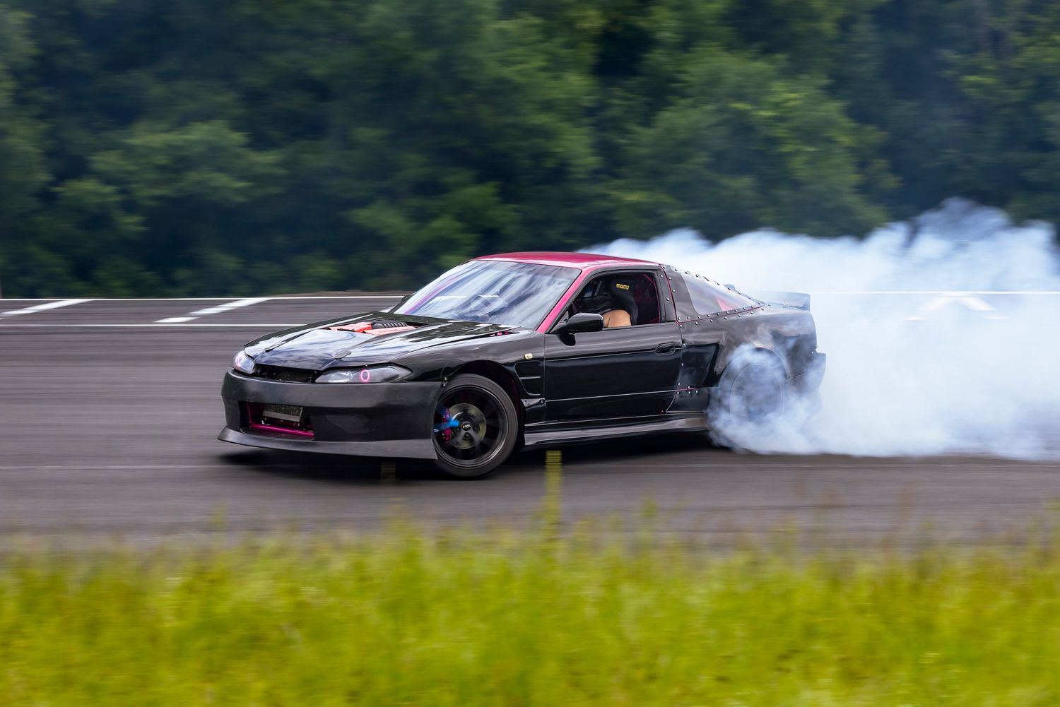 Black drift car with a red-striped hood sliding around a track corner while billowing white tire smoke beside green grass.