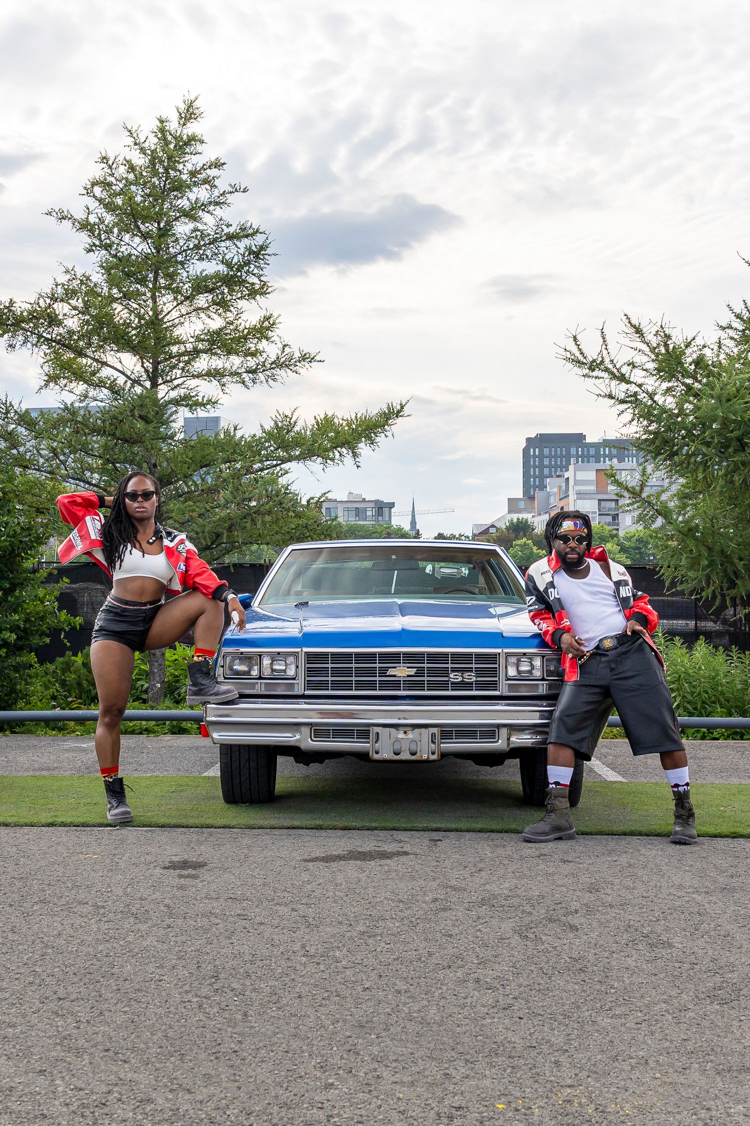 Two people in red jackets and sunglasses pose in front of a blue vintage Chevrolet with trees and city buildings behind.