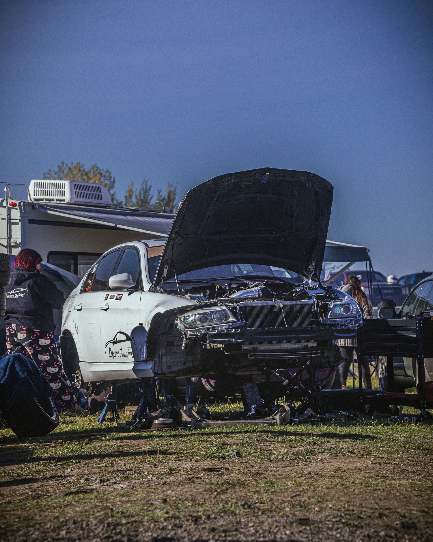 White sports car with its hood up and engine exposed, being worked on in a dusty paddock beside parked trailers.