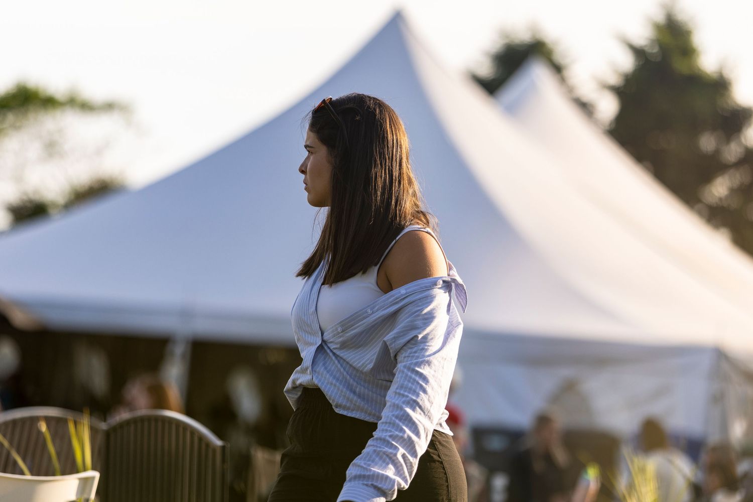 Woman in a white knotted shirt stands outdoors in golden evening light near large white festival tents and seated guests.