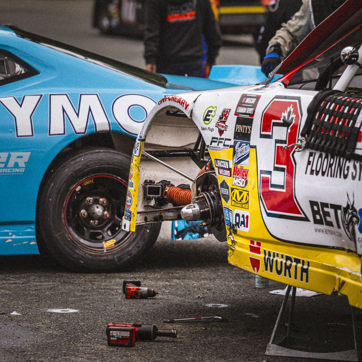 Mechanic using a yellow tape measure to check suspension alignment next to a brake caliper on a race car.