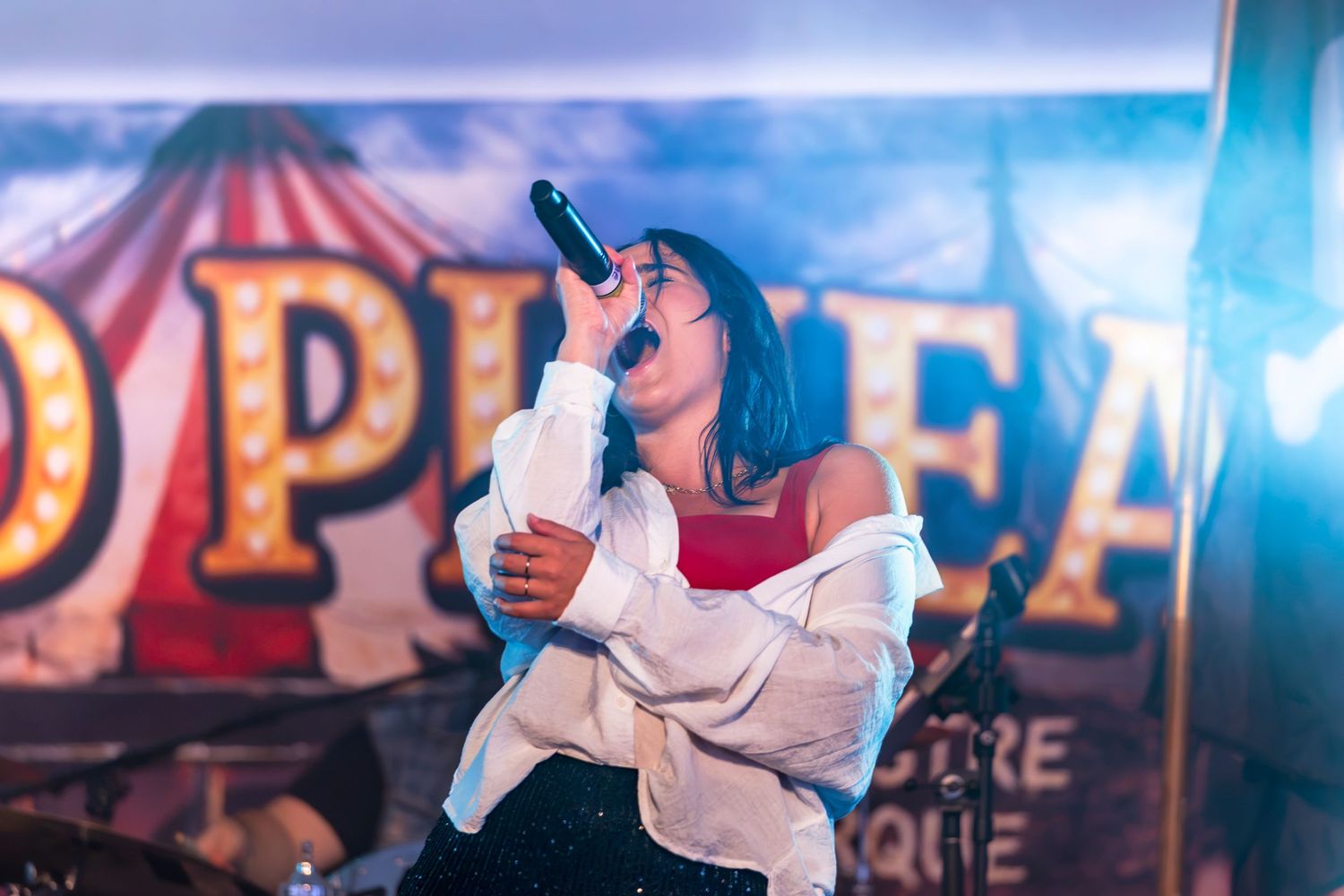 A female singer in a white jacket and red top performs passionately into a microphone on stage in front of a vintage circus-style backdrop.