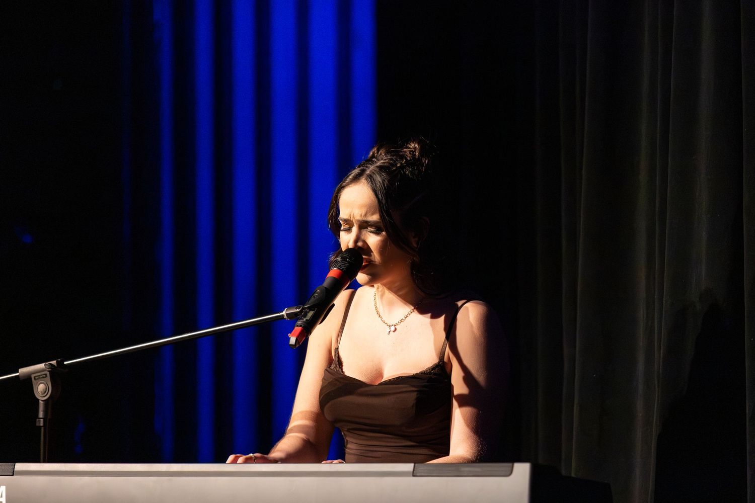 A woman in a black dress sings into a microphone while playing a keyboard on a dark stage with blue curtains.