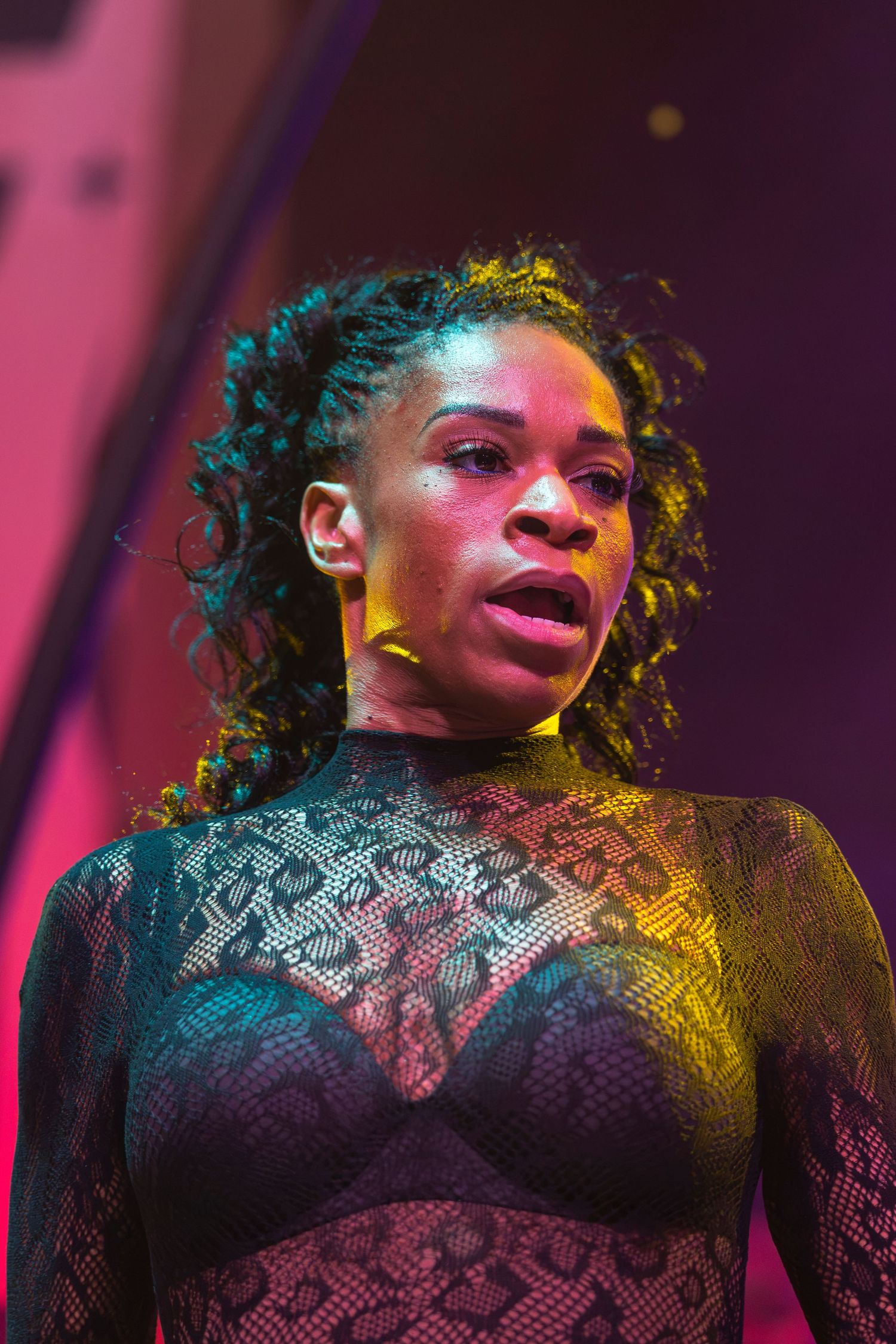 Performer in a black lace bodysuit sings on stage with curly hair and warm yellow and pink concert lighting.