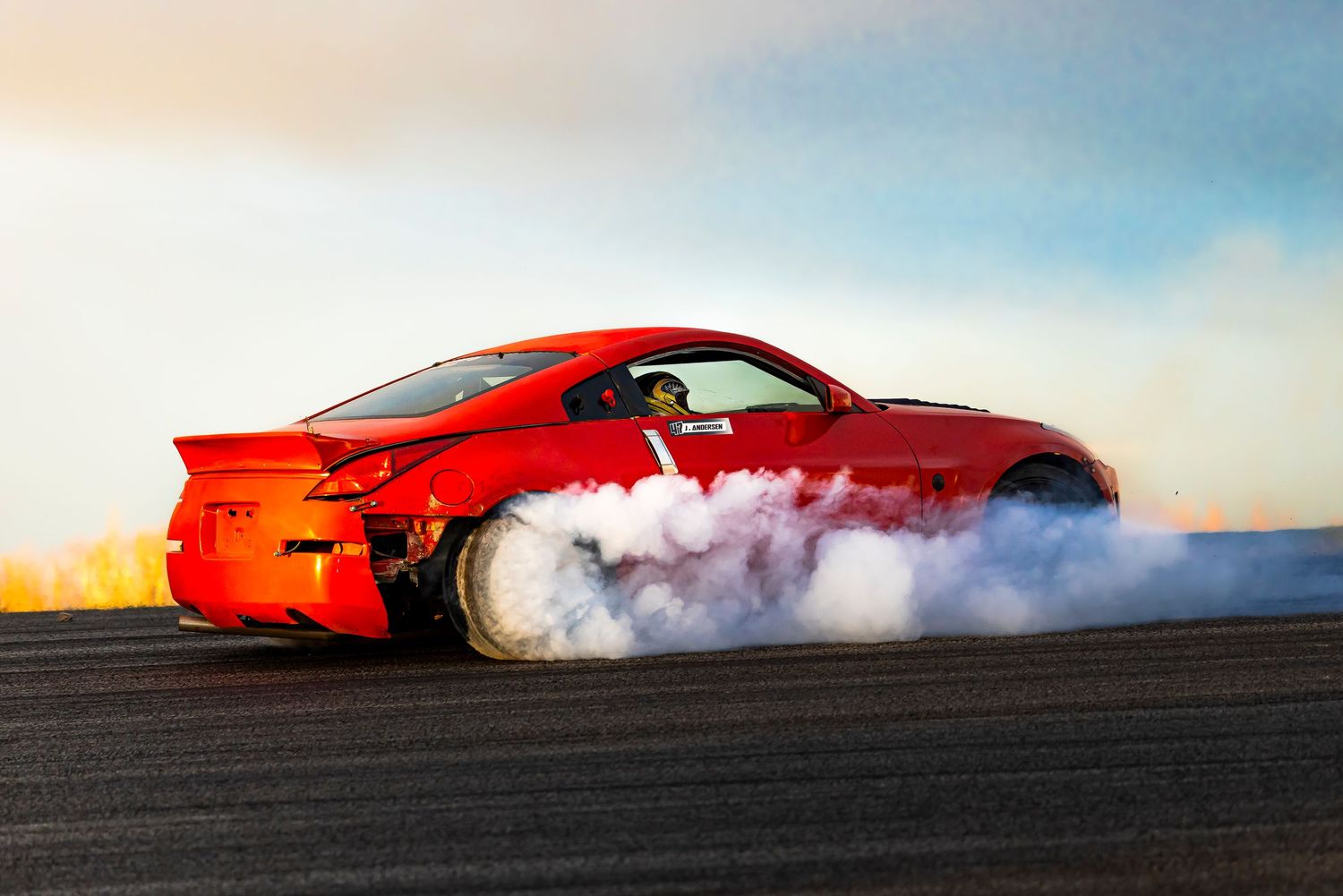 Red sports car drifting around a track at sunset while thick white tire smoke billows from the rear wheel.