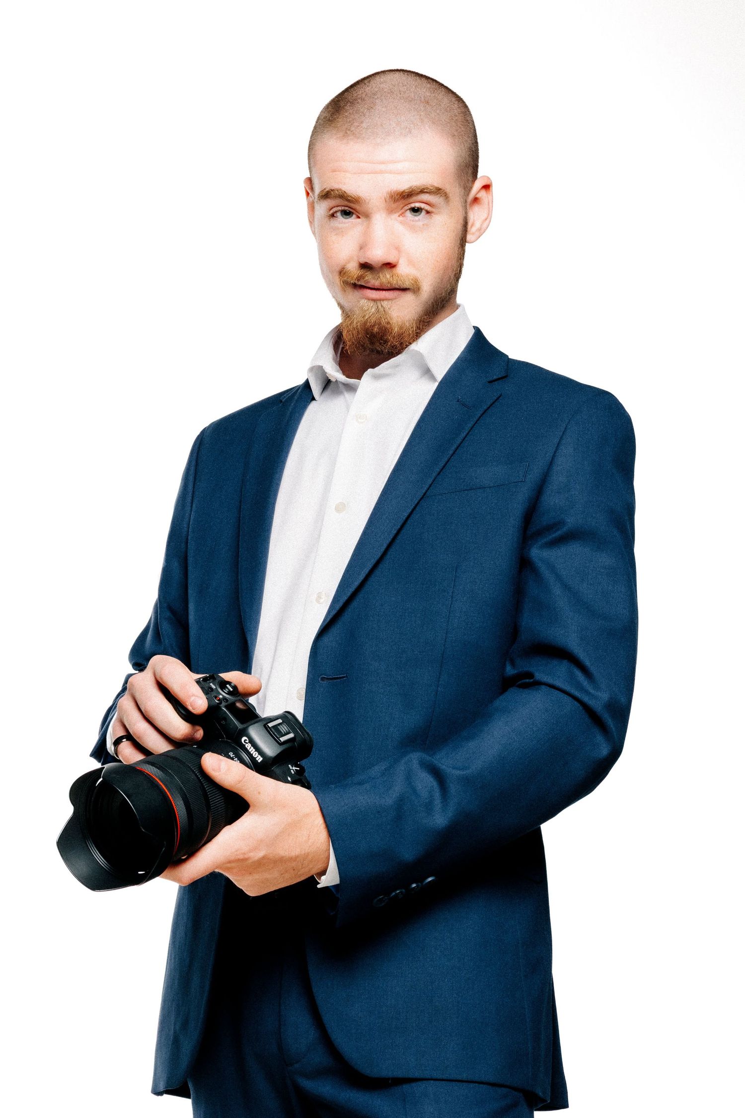 Smiling bald photographer with a red beard wearing a blue suit jacket and holding a DSLR camera in a studio portrait.