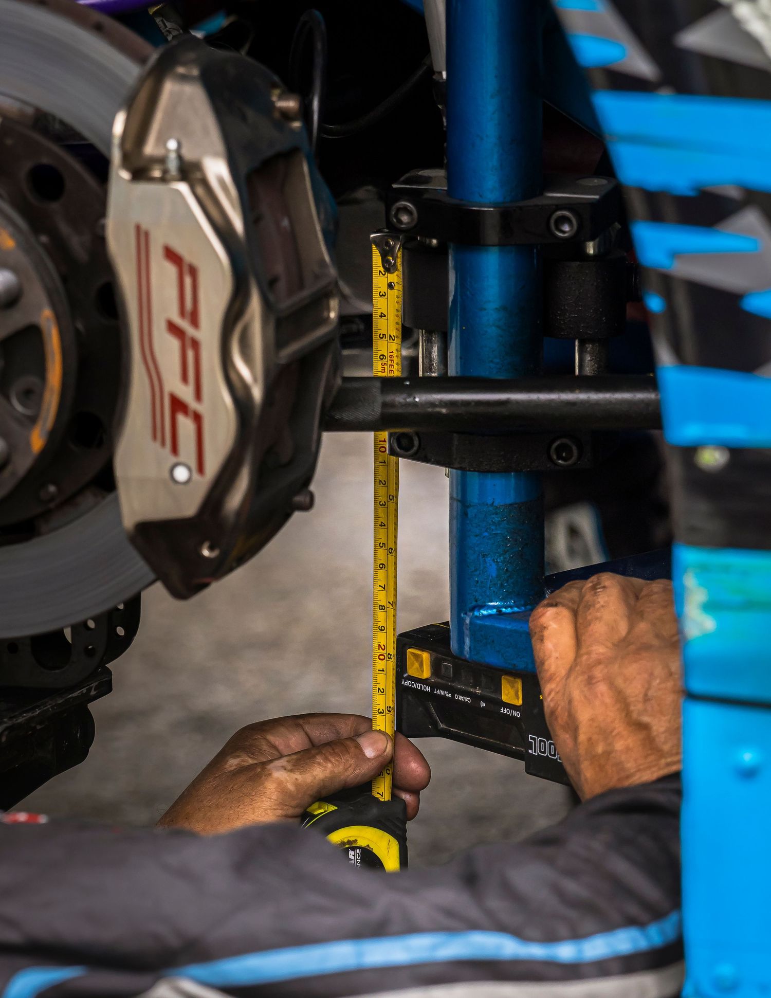 A mechanic's hands use a yellow tape measure on a race car suspension next to a PFC brake caliper and disc.