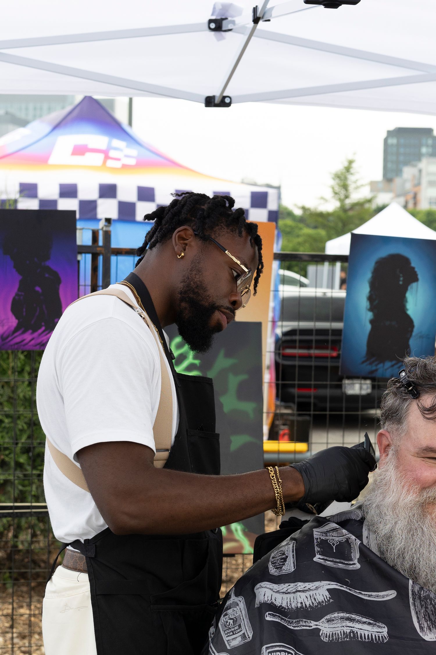 A barber in an apron and glasses trims a bearded client's hair under a white tent at an outdoor event.