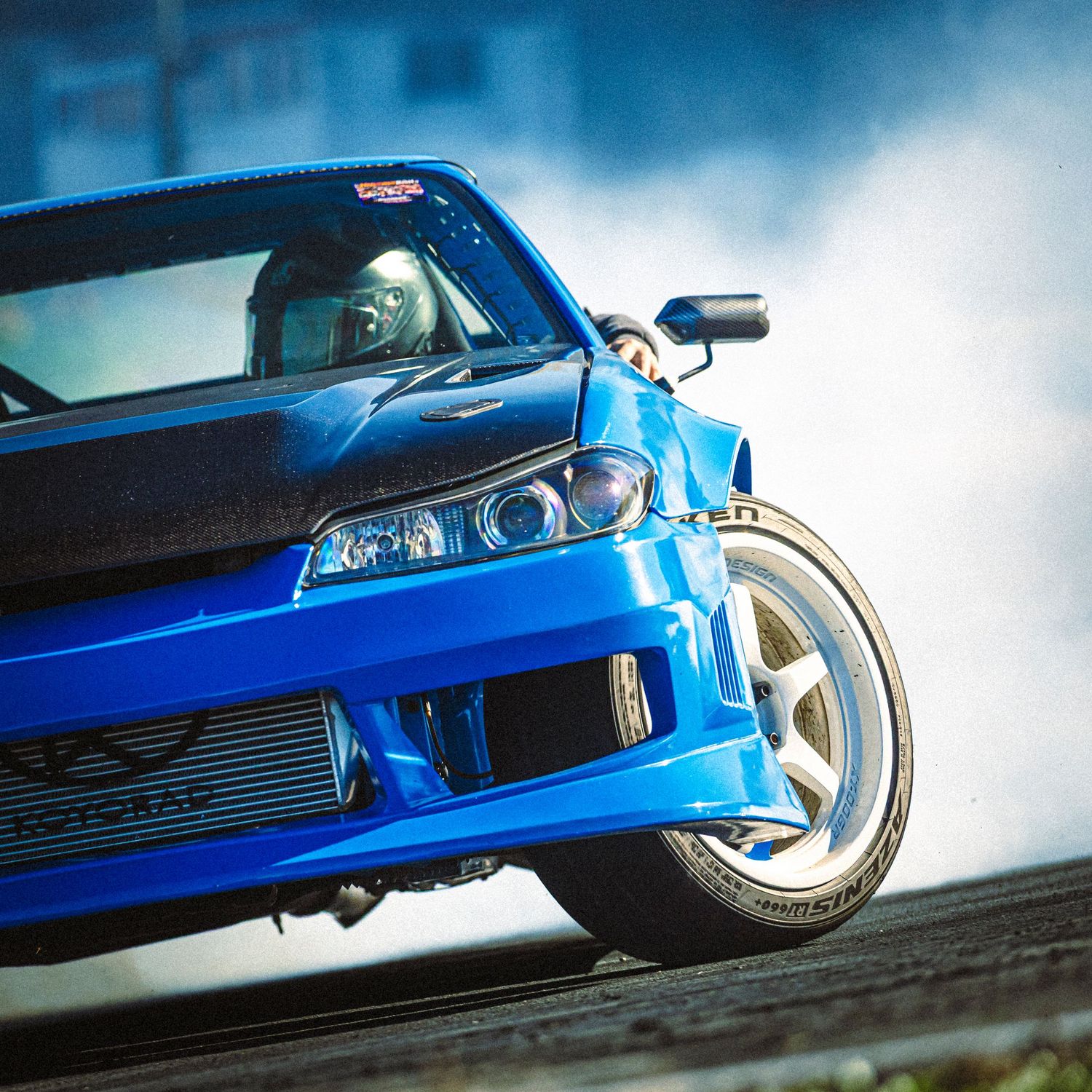Close-up of a blue drift car's front end with a helmeted driver visible as the car turns on a smoky track.